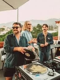 Three people at a social gathering outdoors, with a DJ table and equipment, under an umbrella, and scenic hills in the background.