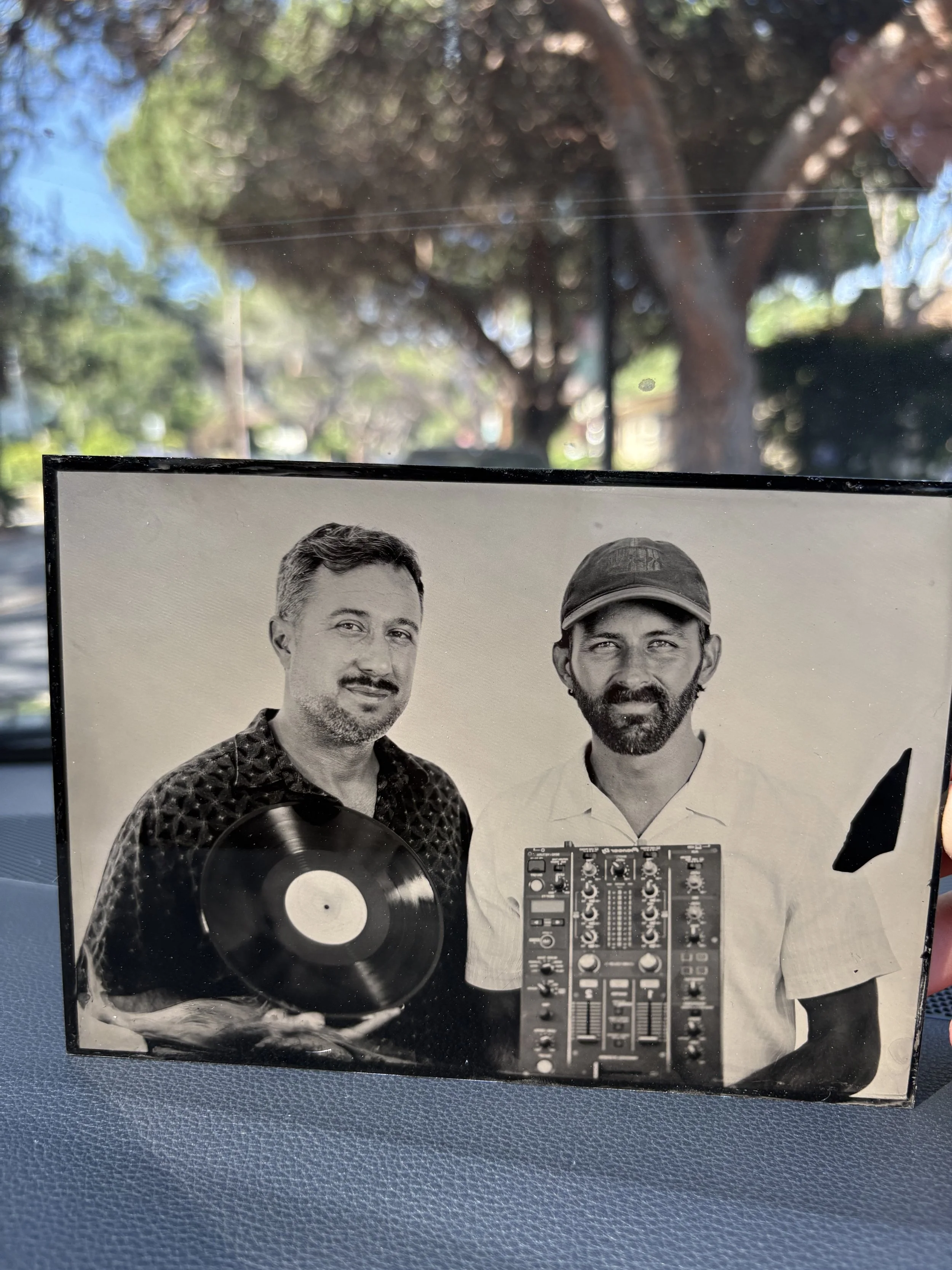 Black and white photo of two men, one with short hair and a patterned shirt, the other with a beard and cap, standing behind a DJ mixer and a vinyl record.