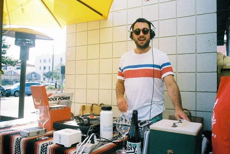 DJ wearing headphones and sunglasses DJing at an outdoor event with a table of equipment, drink, and a yellow umbrella in the background.