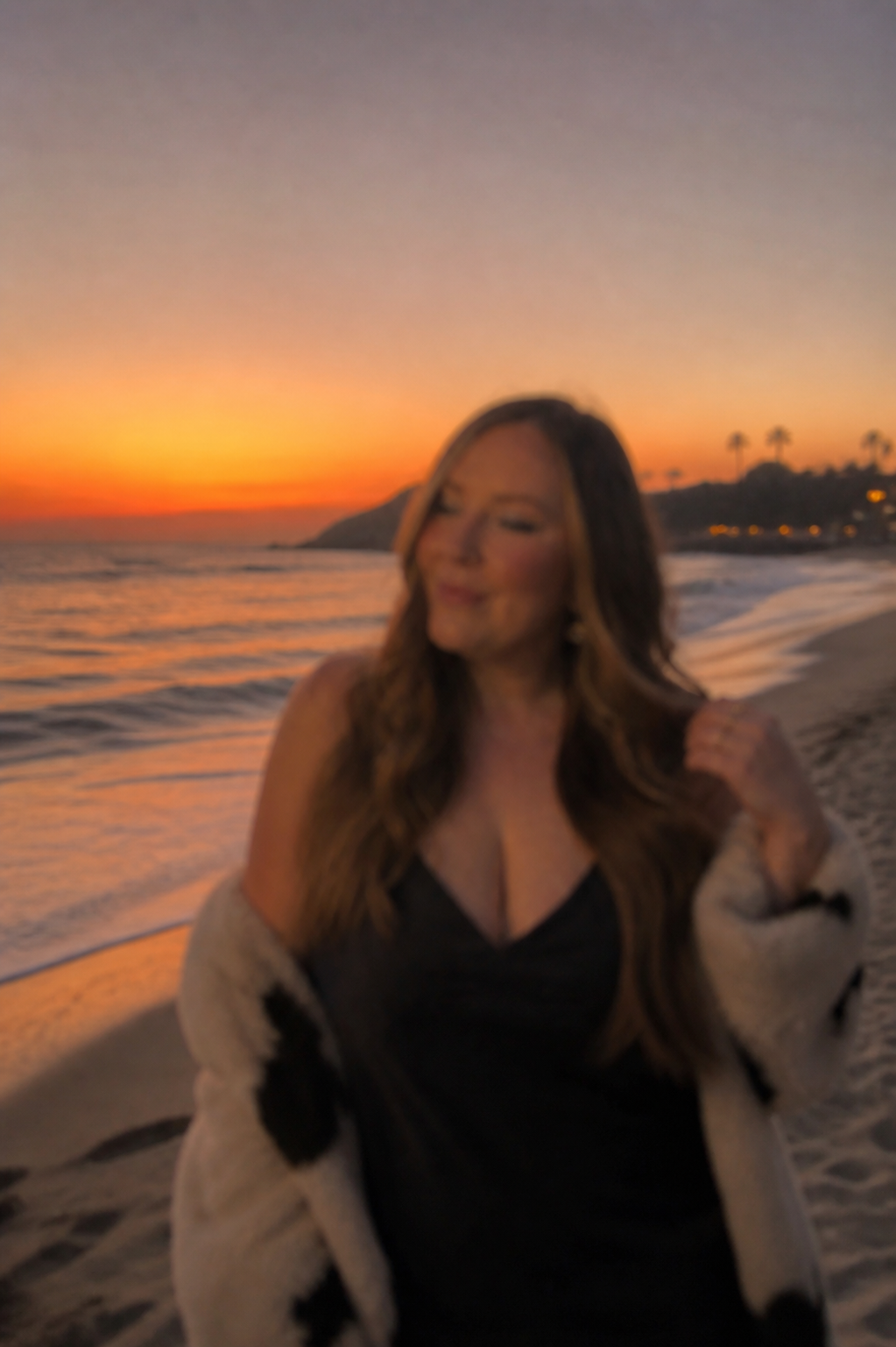 A woman on a beach at sunset, with palm trees and silhouette hills in the background, smiling and wearing a black top and a cream-colored cardigan with black spots.