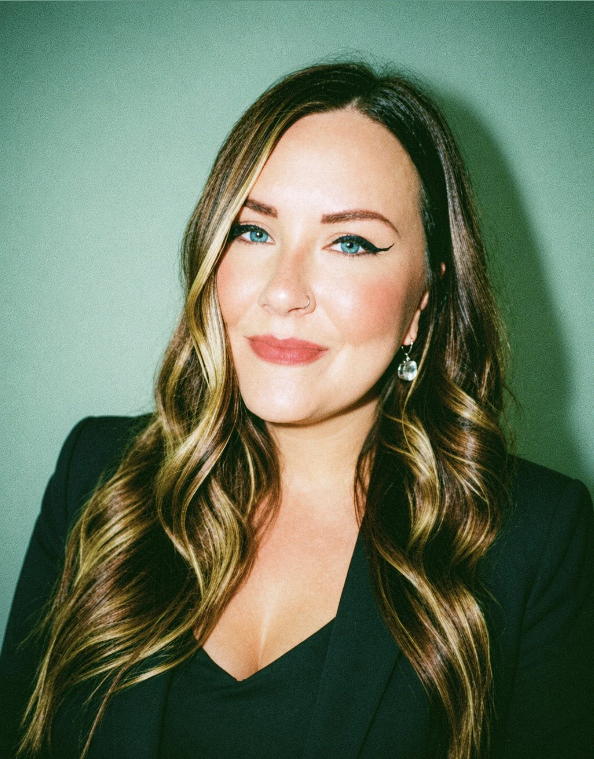 A woman with long, wavy brown hair streaked with blonde highlights, wearing a black top and black blazer, smiling softly and looking at the camera. She has bright blue eyes, a nose piercing, and a dangling earring, against a plain light green background.