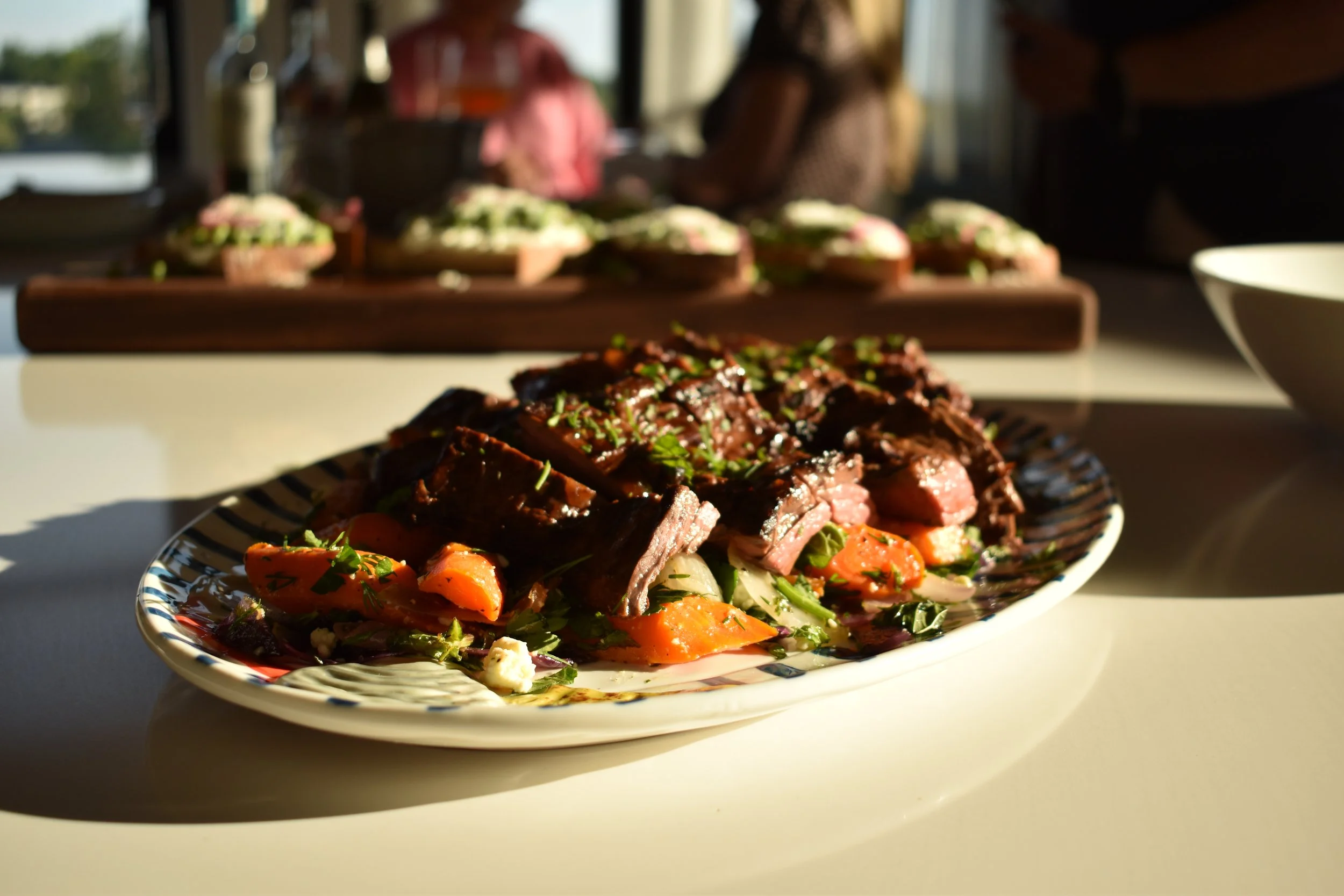 A plate of cooked beef with herbs and vegetables, in front of a blurry background of people preparing food.