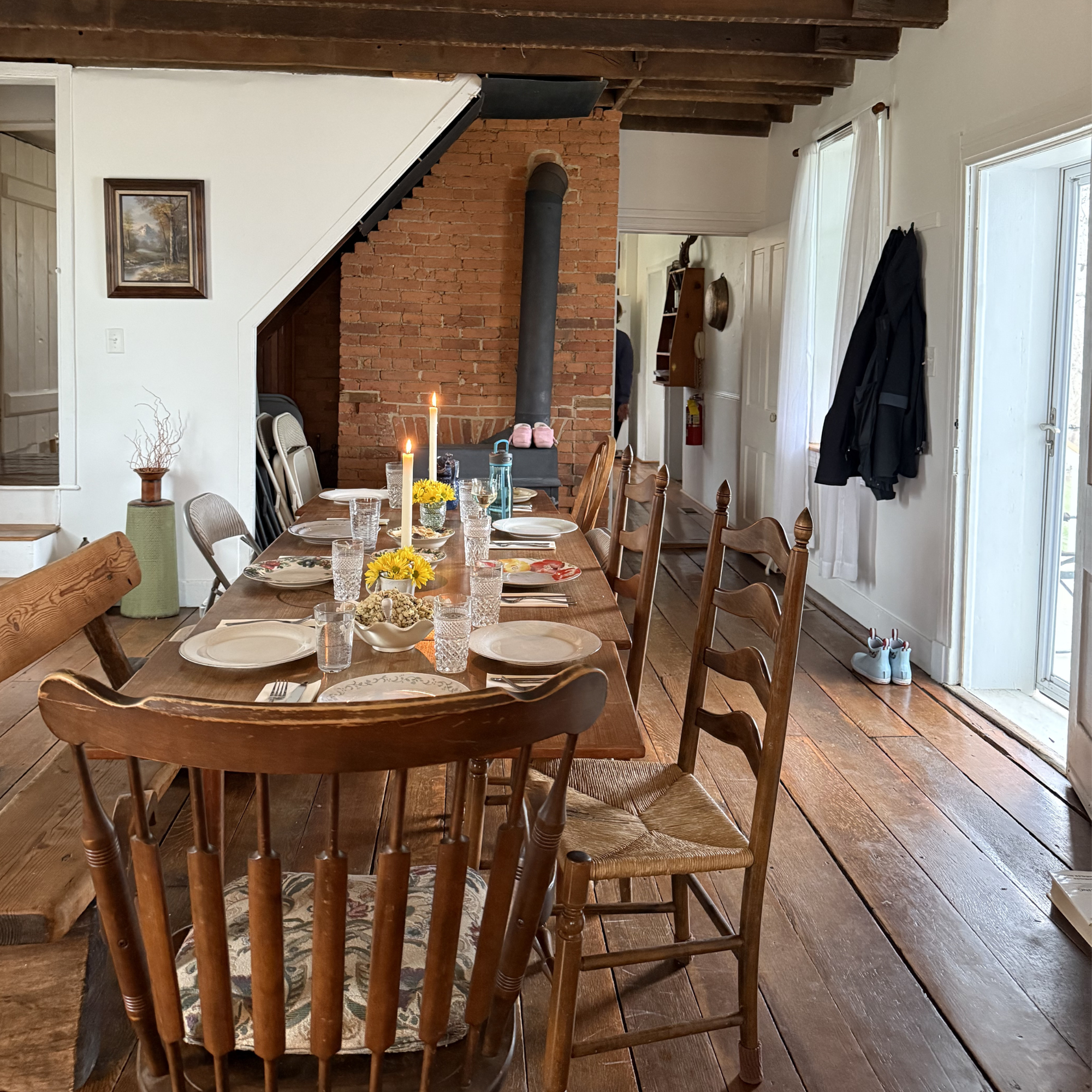 A wooden dining table set for a meal with white plates, drinking glasses, candles, and a small vase with yellow flowers, in a rustic dining room with wood floors and white walls, featuring a staircase and a brick fireplace.