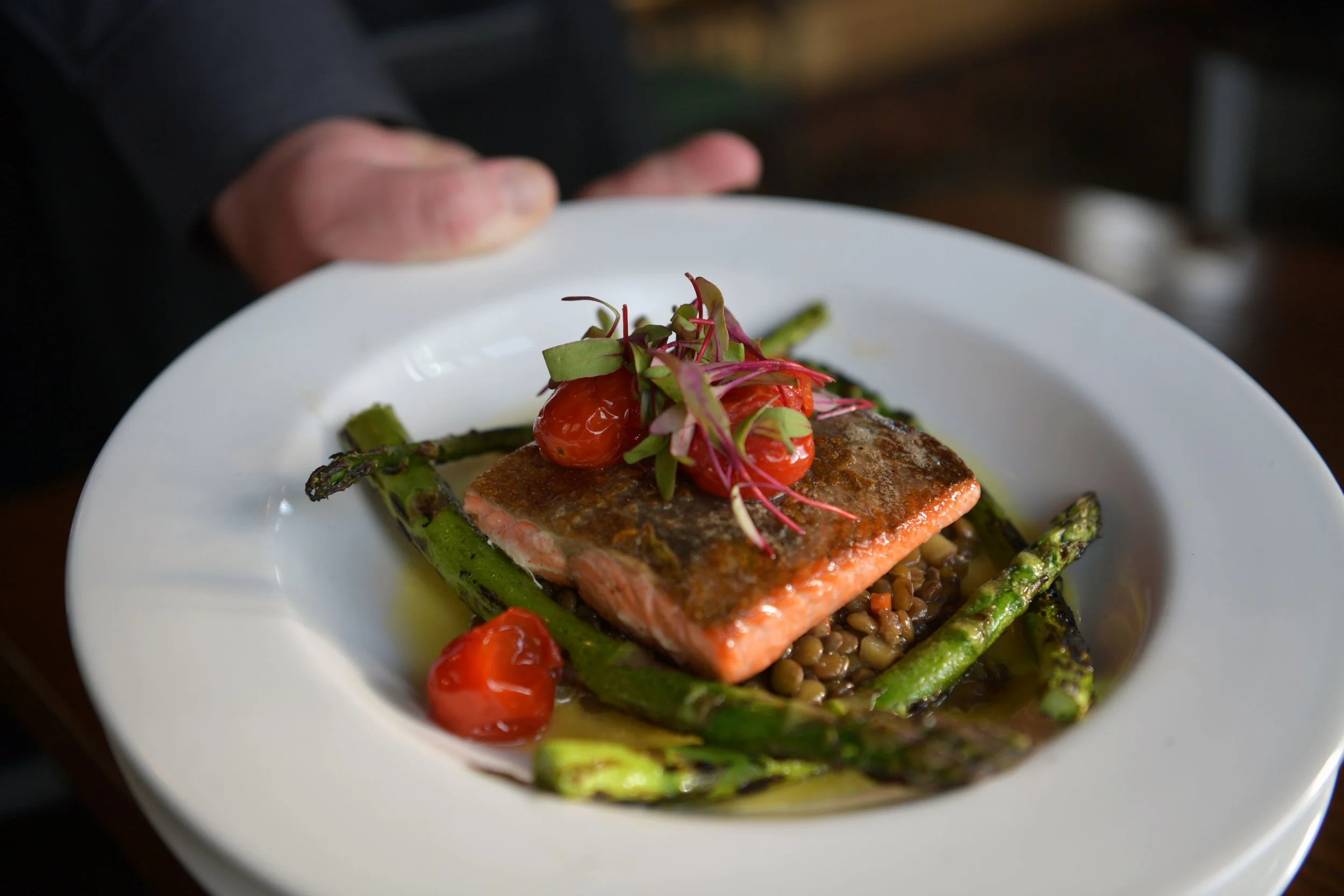 A plate of cooked salmon topped with microgreens, cherry tomatoes, and grilled asparagus, served over lentils.
