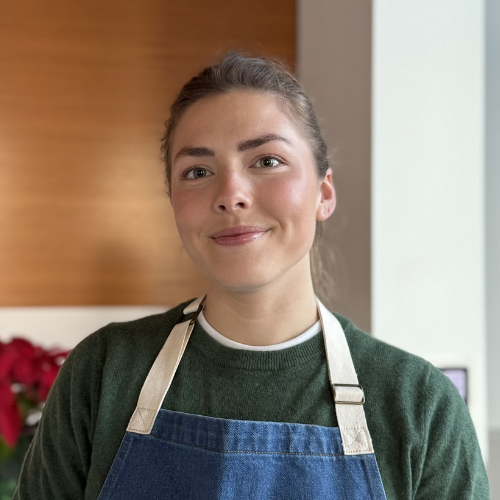 A young woman with brown hair tied back, wearing a green sweater and a blue apron, smiling in a kitchen with wooden and white walls.