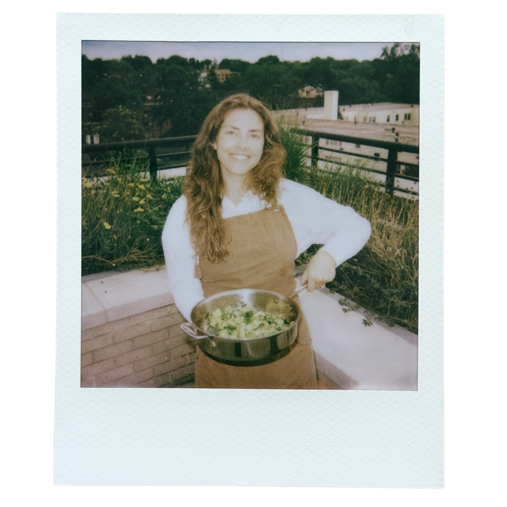 A smiling woman with long, curly hair stands on a rooftop garden with a large salad bowl filled with greens in front of her. In the background, there are trees, buildings, and a cloudy sky.