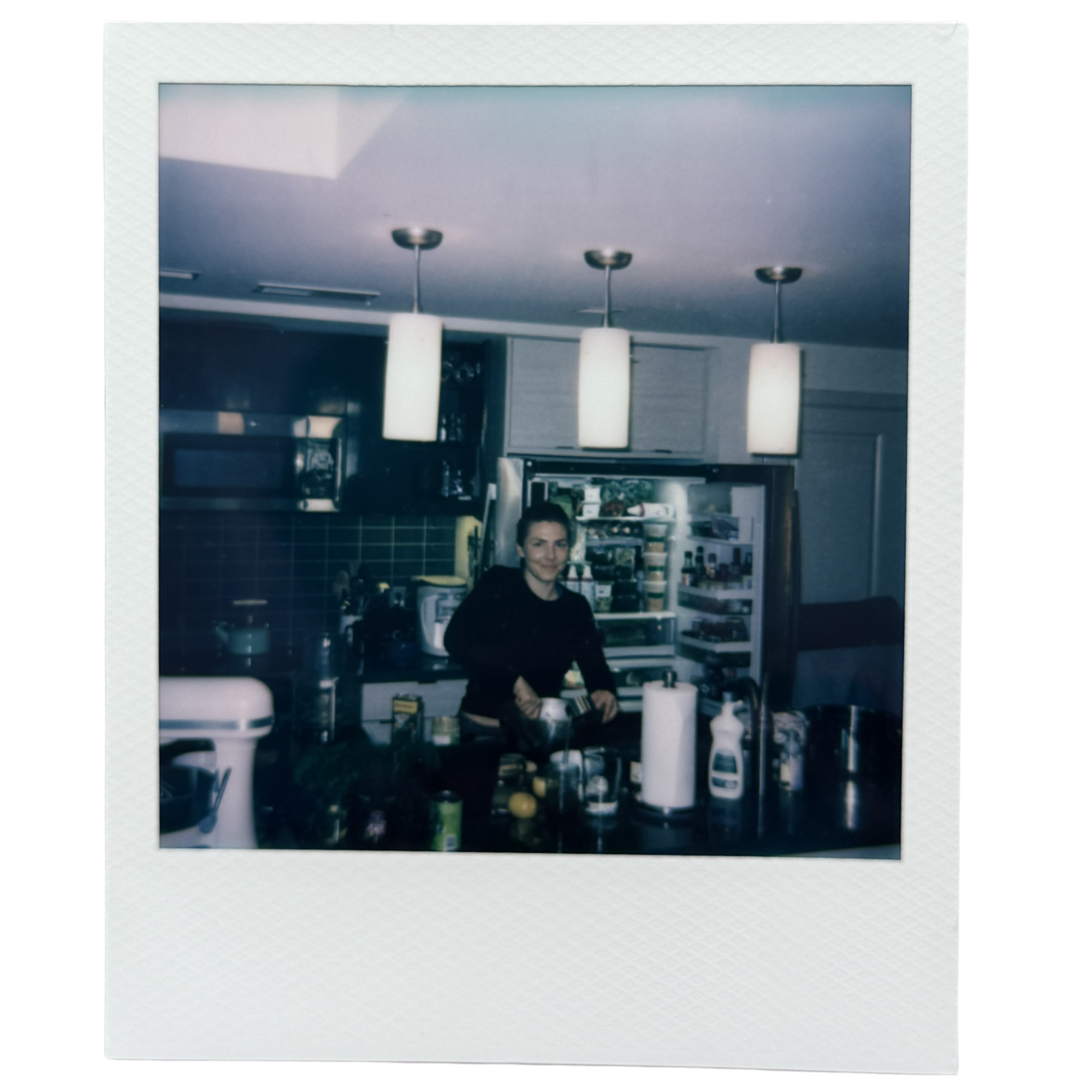 Polaroid style photograph of a woman with short hair sitting at a table in a modern interior space, with hanging pendant lights and shelves with items in the background.