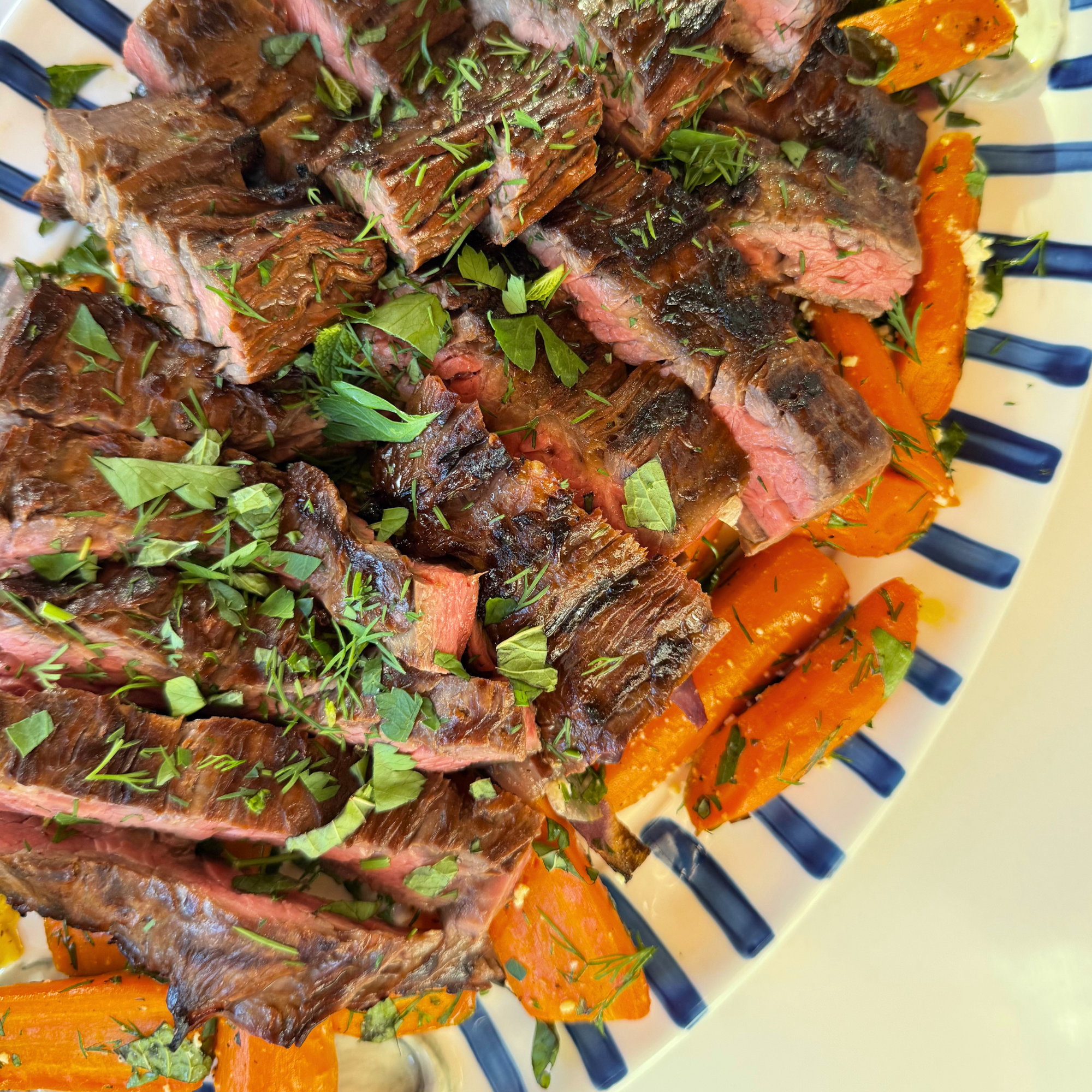 Slices of cooked steak garnished with chopped green herbs, served with roasted carrots on a striped plate.