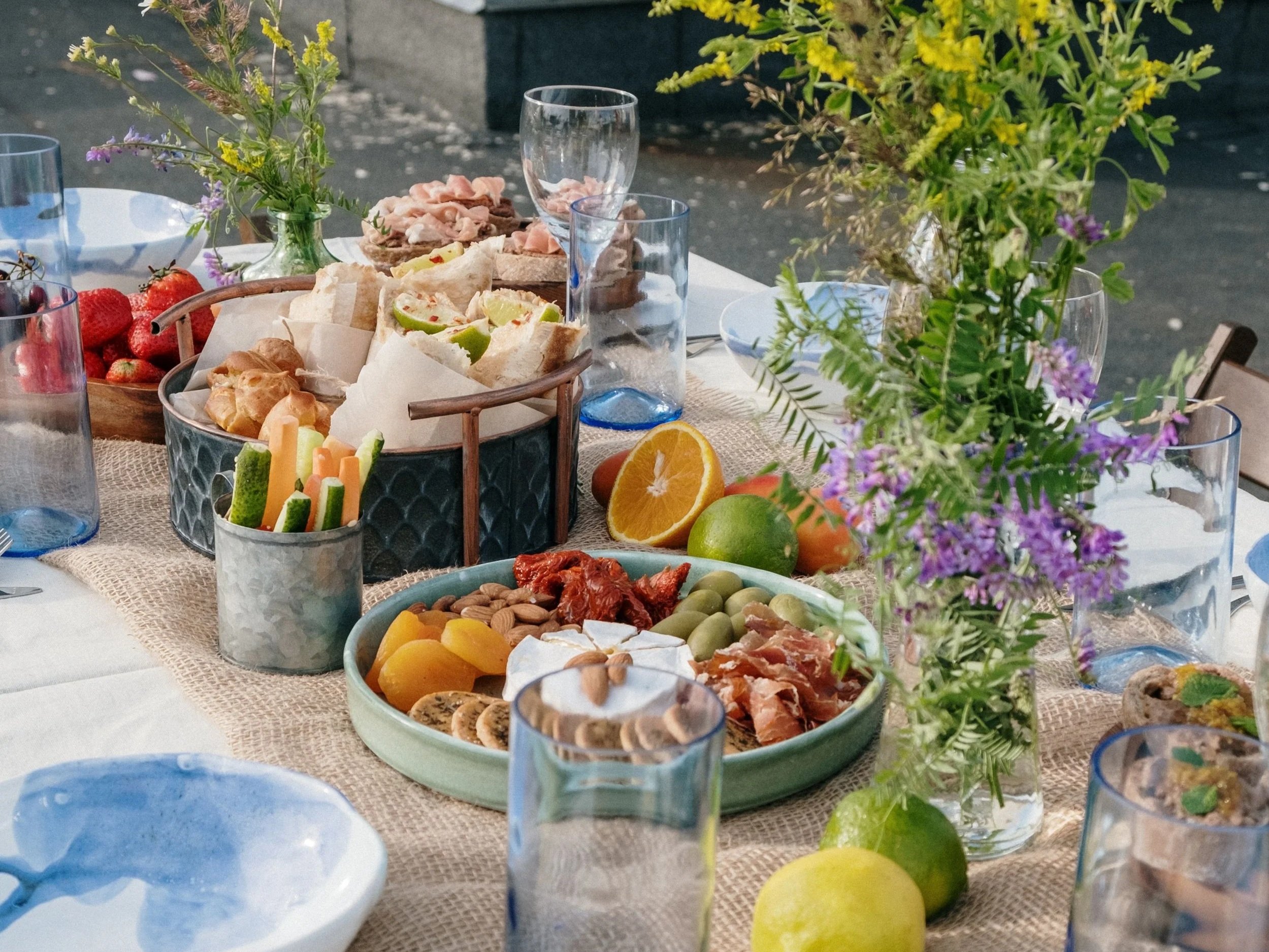 A table set with various bowls and plates of food, including fruits, vegetables, cheeses, and appetizers, with a centerpiece of colorful flowers.