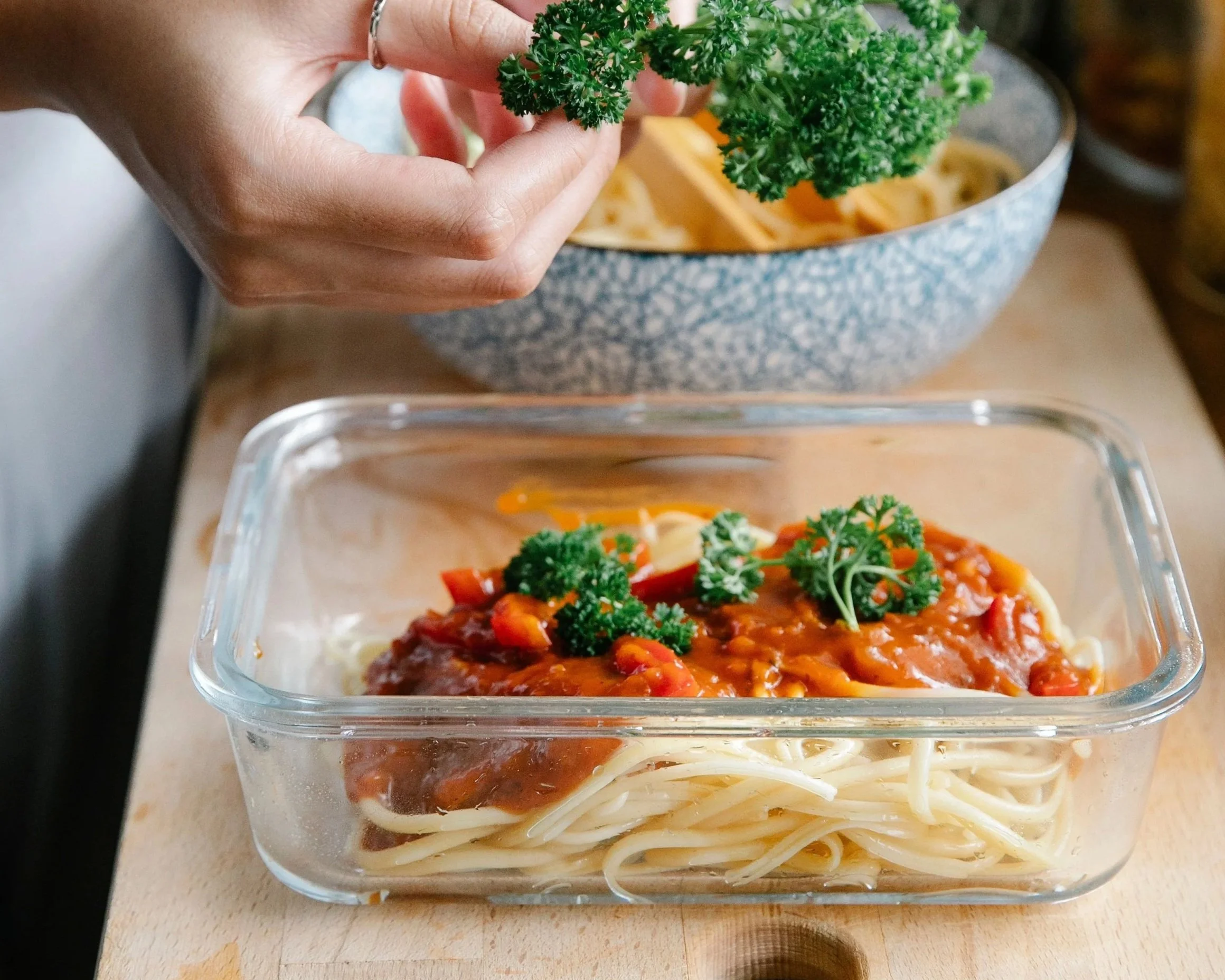 A hand holding fresh parsley over a dish with spaghetti noodles topped with meat sauce and garnished with parsley, in a glass baking dish.