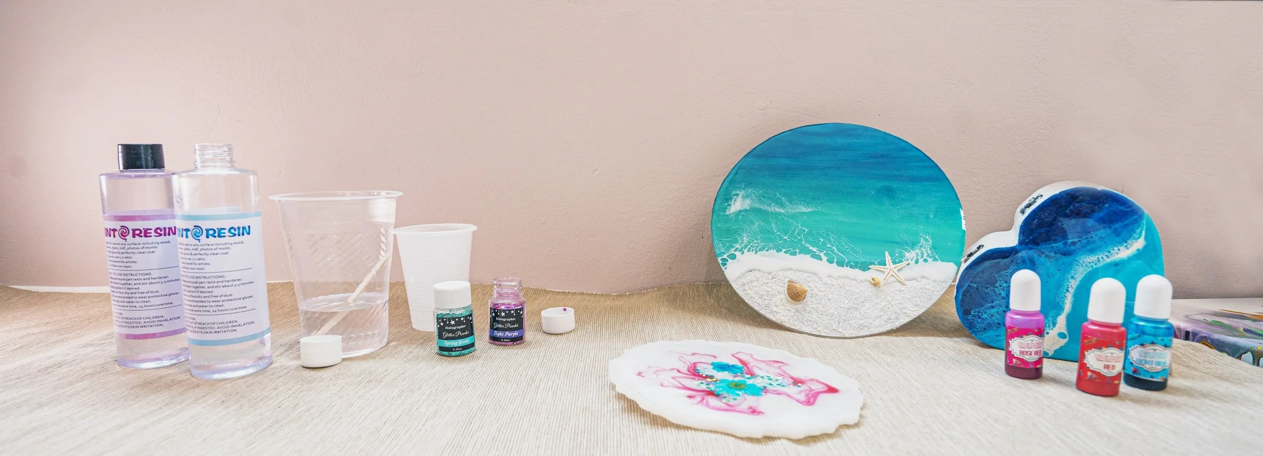 Surfboard-shaped art piece with beach scene, starfish, and shells, alongside resin art supplies like bottles of resin, colored pigments, small cups, and a decorated resin piece on a table.