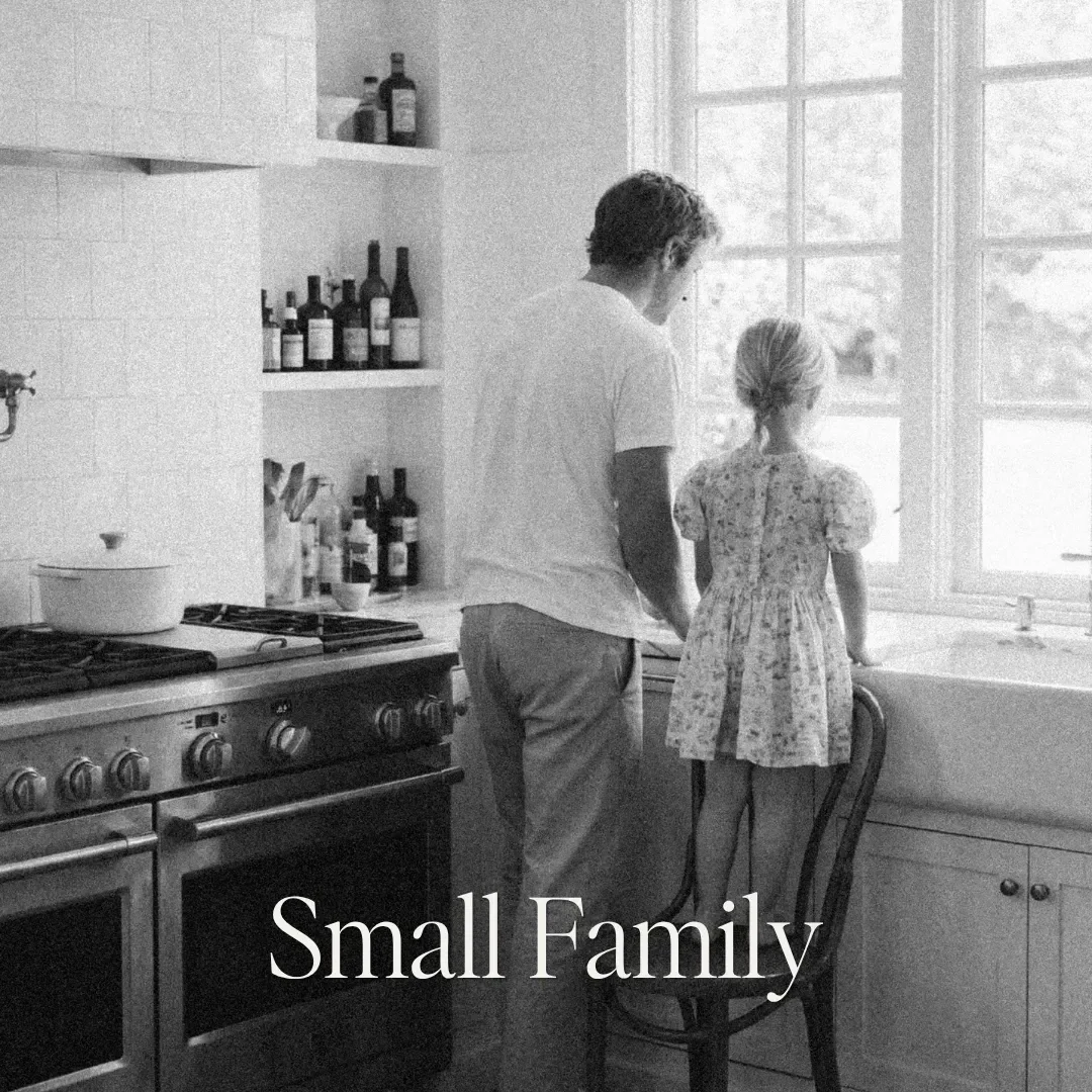 A man and a young girl standing by a kitchen window. The girl is looking outside, while the man looks at her. The kitchen has open shelves with bottles, a stove, and a pot. The text 'Small Family' is overlaid on the image.
