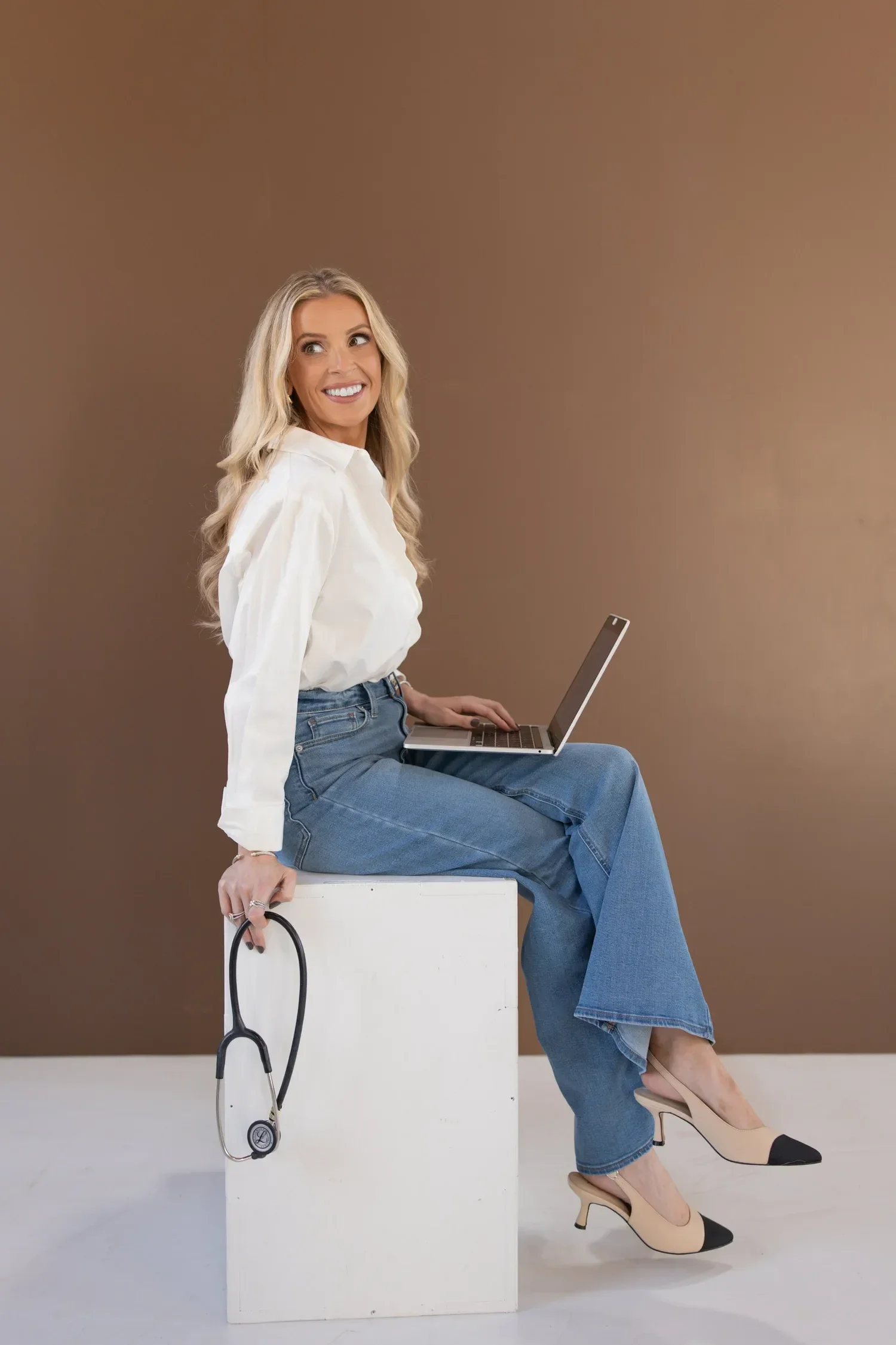 Dr. Maurine Hart in a white blouse and blue jeans sitting cross-legged on a white box, holding a stethoscope in her right hand and a laptop on her lap, against a plain brown background.