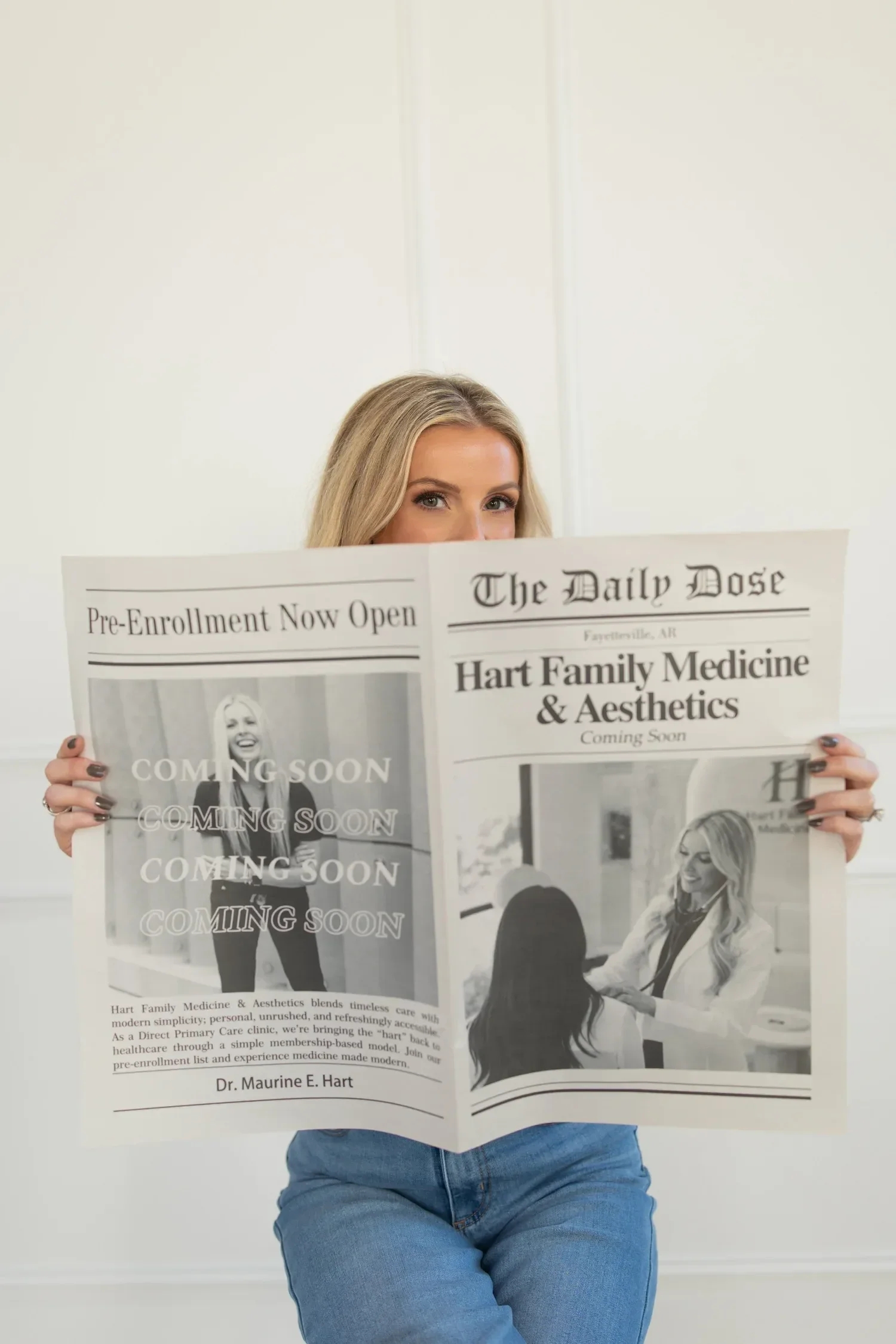 Dr. Maurine Hart sitting with crossed arms and blue jeans, holding a newspaper in front of their face that says 'COMING SOON' and features pictures and text about Hart Family Medicine & Aesthetics.
