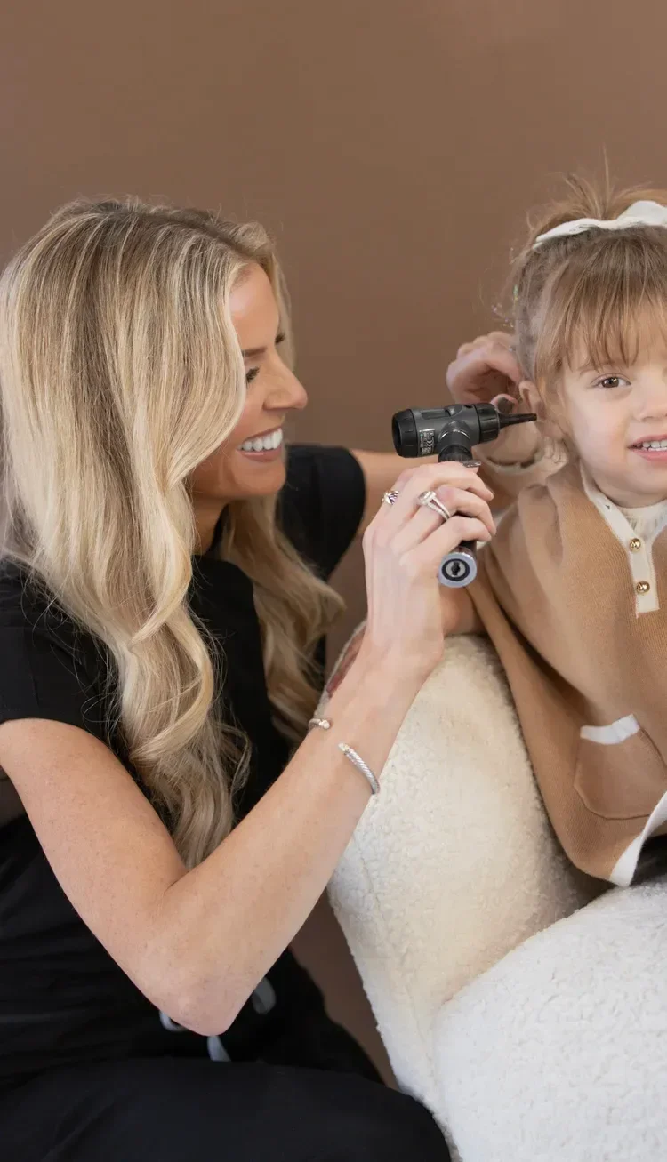 Dr. Maurine Hart smiling as she holds an instrument checking a young girl's right ear with light brown hair, wearing a beige sweater, as they sit in a cozy room.