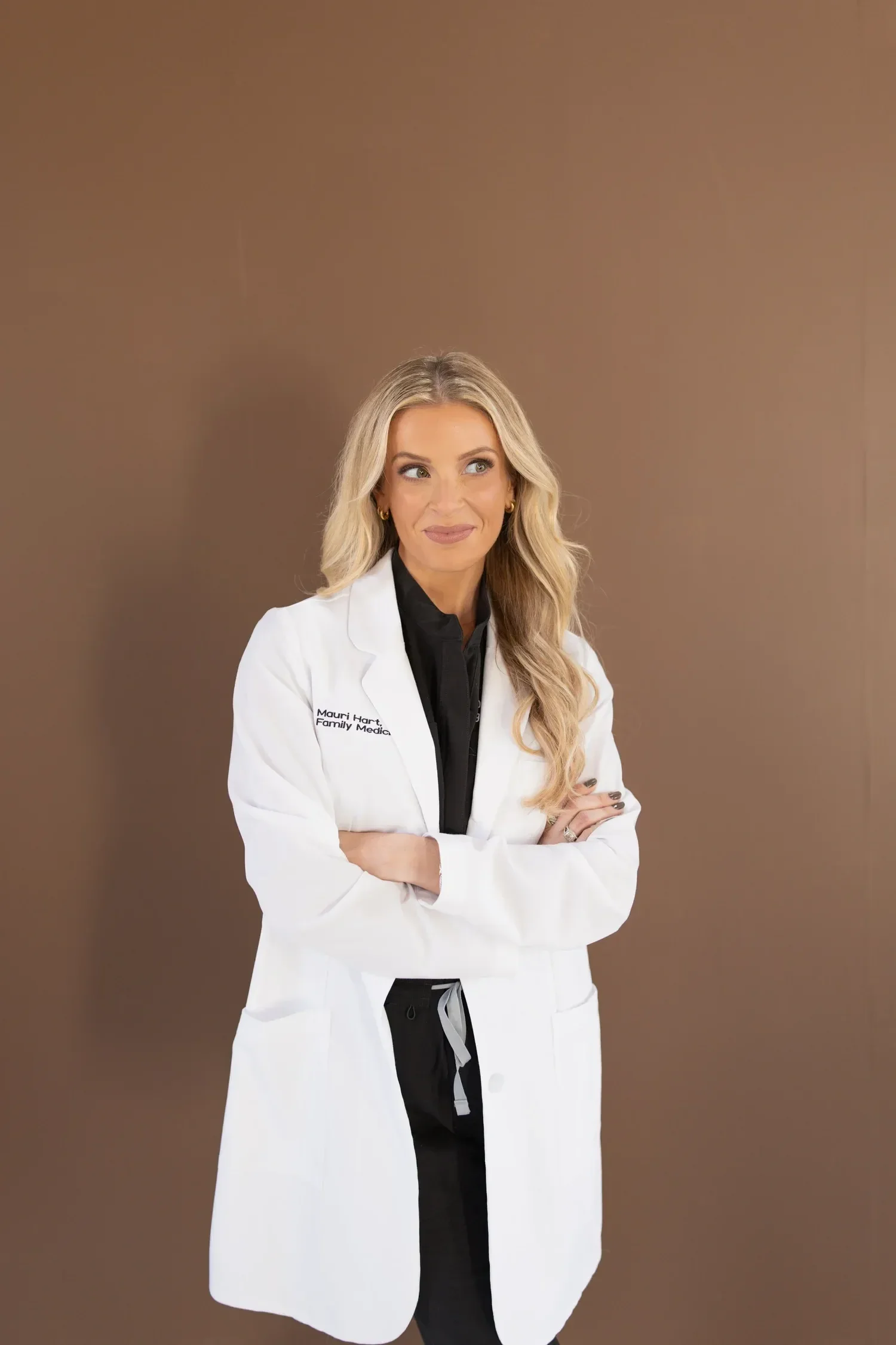 Dr. Maurine Hart with long blond hair, wearing a white medical coat with her arms crossed, standing against a plain brown background.