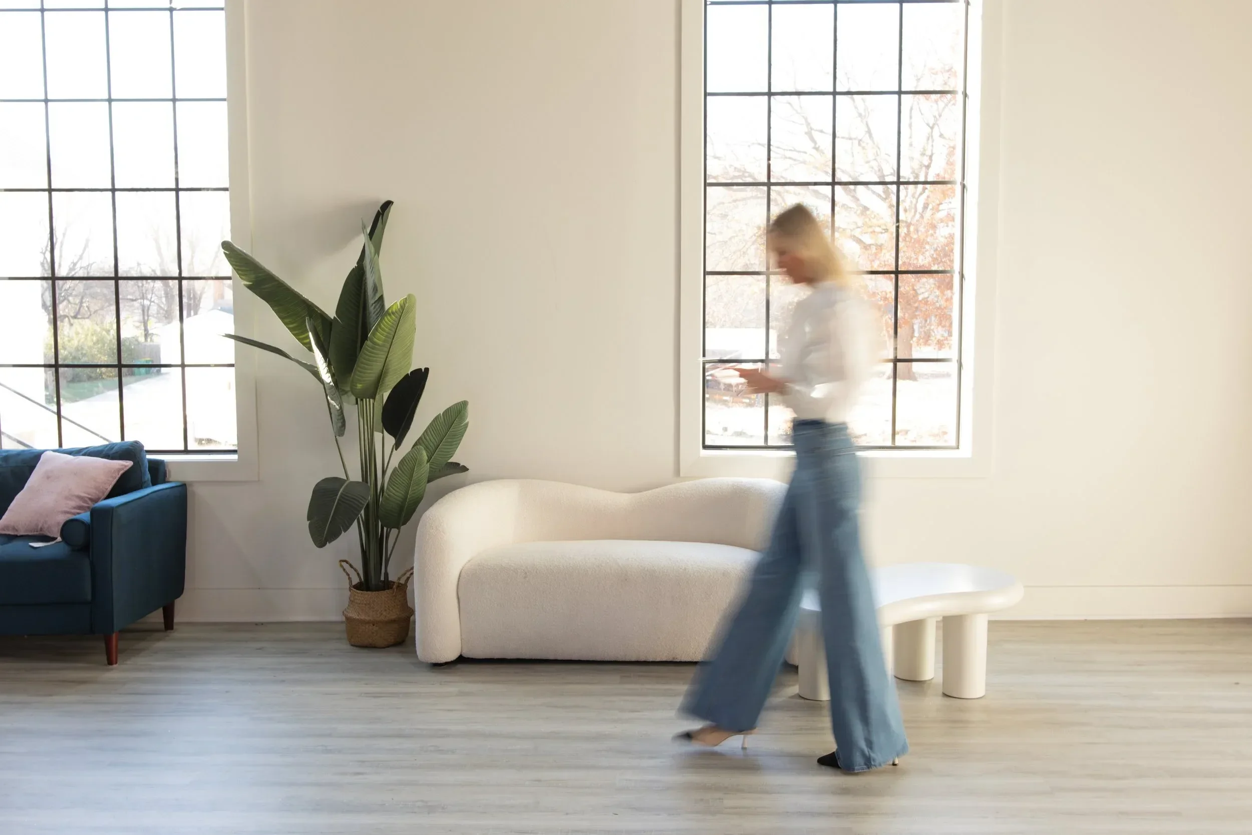 Dr. Maurine Hart walking past large windows in a bright, minimalist living room with a white sofa, a blue armchair, and a tall potted plant.