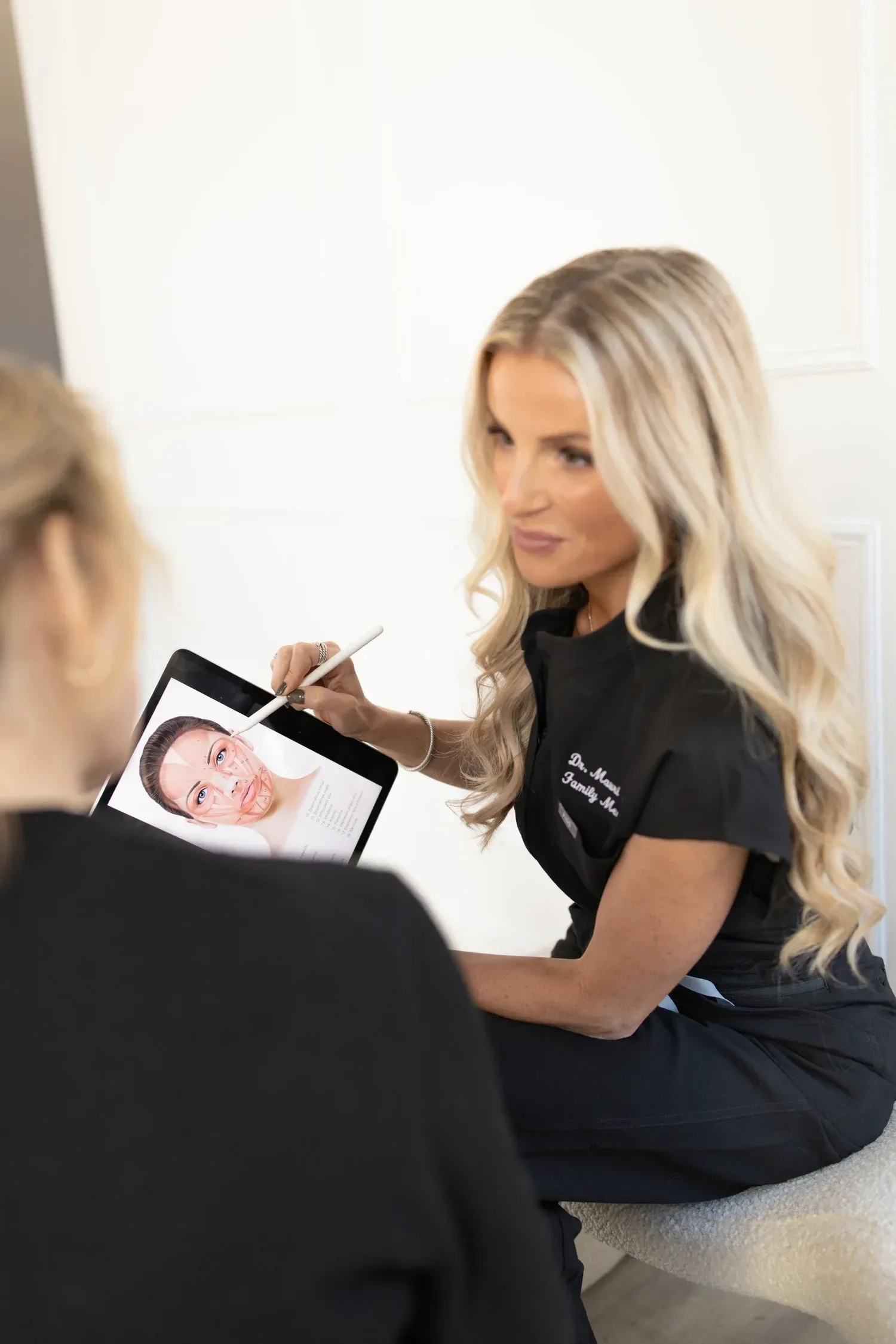 Dr. Maurine Hart wearing a black uniform, is showing a face chart on a tablet to a person in front of her.