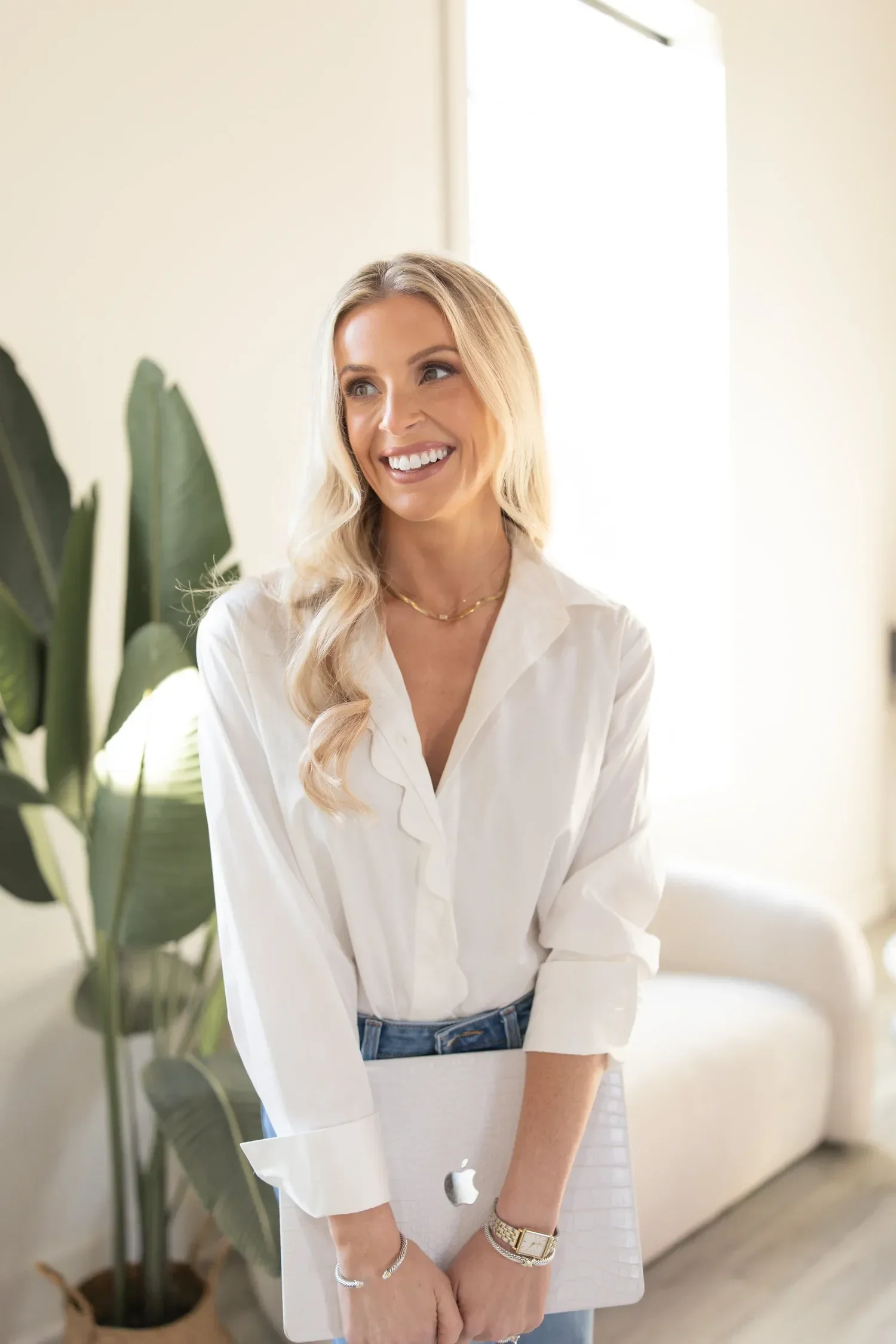 Dr. Maurine Hart with long blonde hair, wearing a white blouse and blue jeans, standing indoors with a large green plant nearby, smiling and holding an Apple iMac computer.