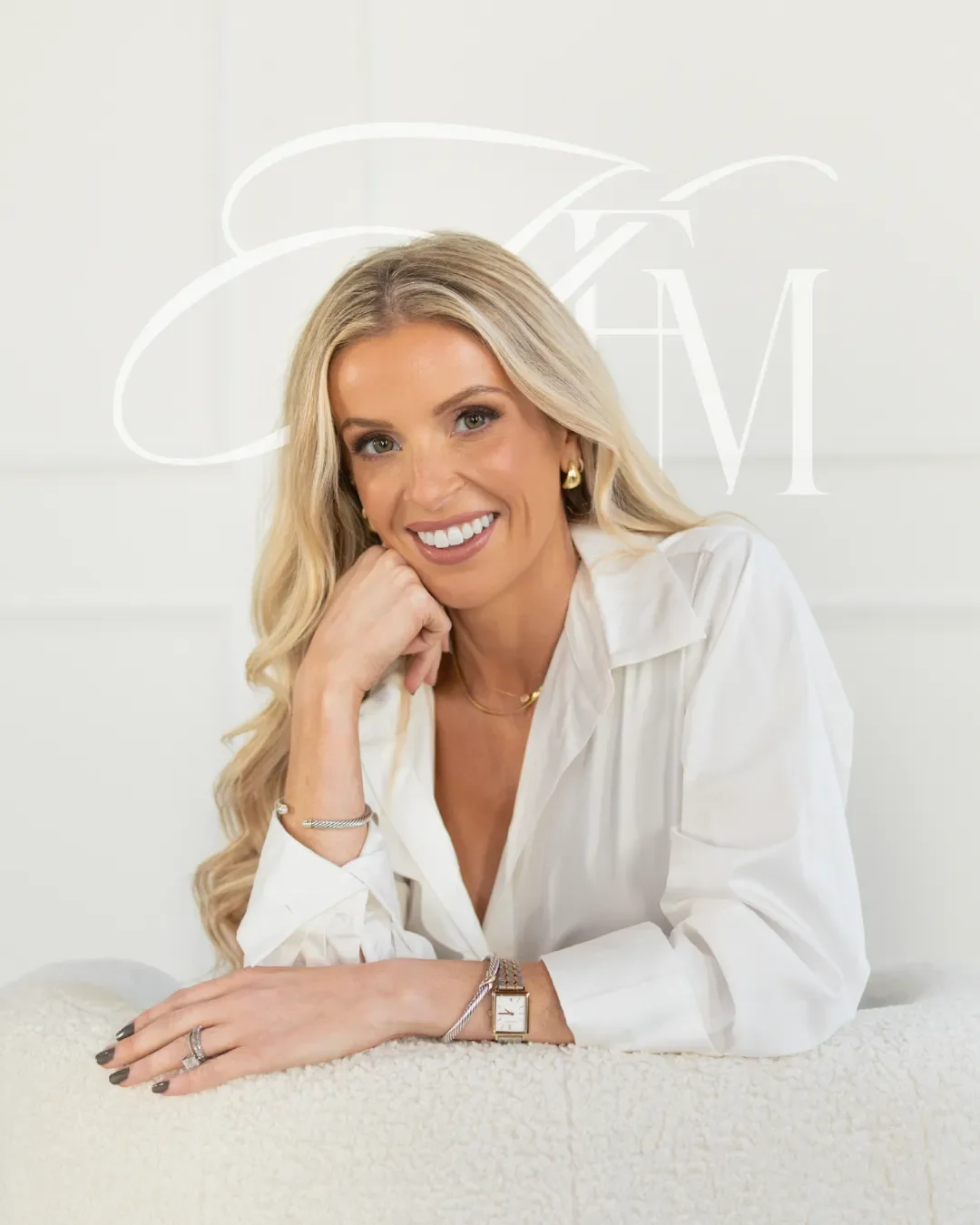 Dr. Maurine Hart with long blonde hair, wearing a white button-up shirt and gold jewelry, resting her chin on one hand, sitting on a white surface with a light background and a logo behind her.