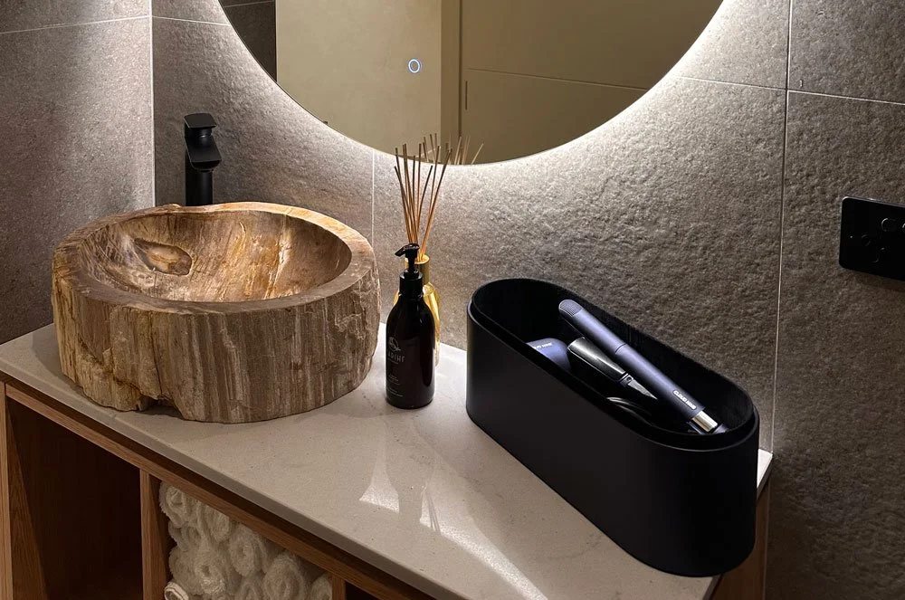 Bathroom vanity with a rustic wooden sink, black faucet, reed diffuser, and a black container holding grooming tools, reflected in a circular mirror on a textured wall.