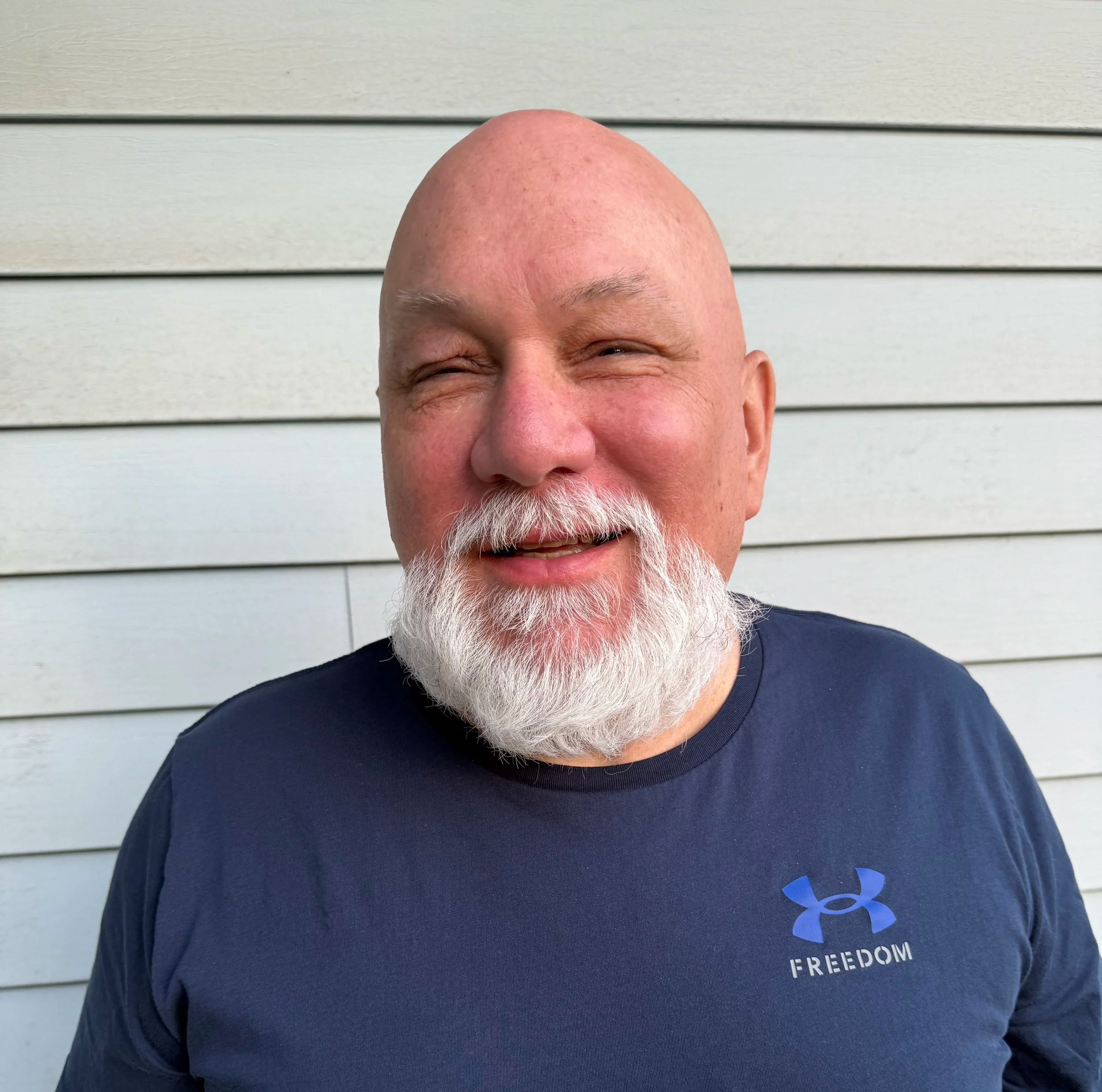 Close-up of a smiling, bald man with a white beard, wearing a dark Under Armour T-shirt with the word 'FREEDOM' visible on it, standing in front of horizontal light gray siding.