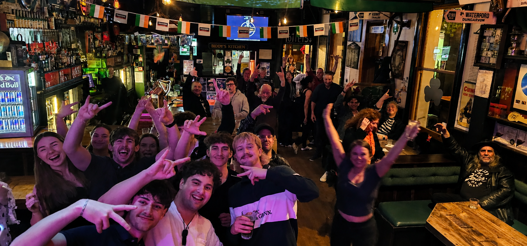 A lively bar scene with smiling, excited people enjoying drinks and dancing. The bar is decorated with Irish flags and various bottles and signage, with a group gathered near the counter and others dancing and celebrating throughout the space.