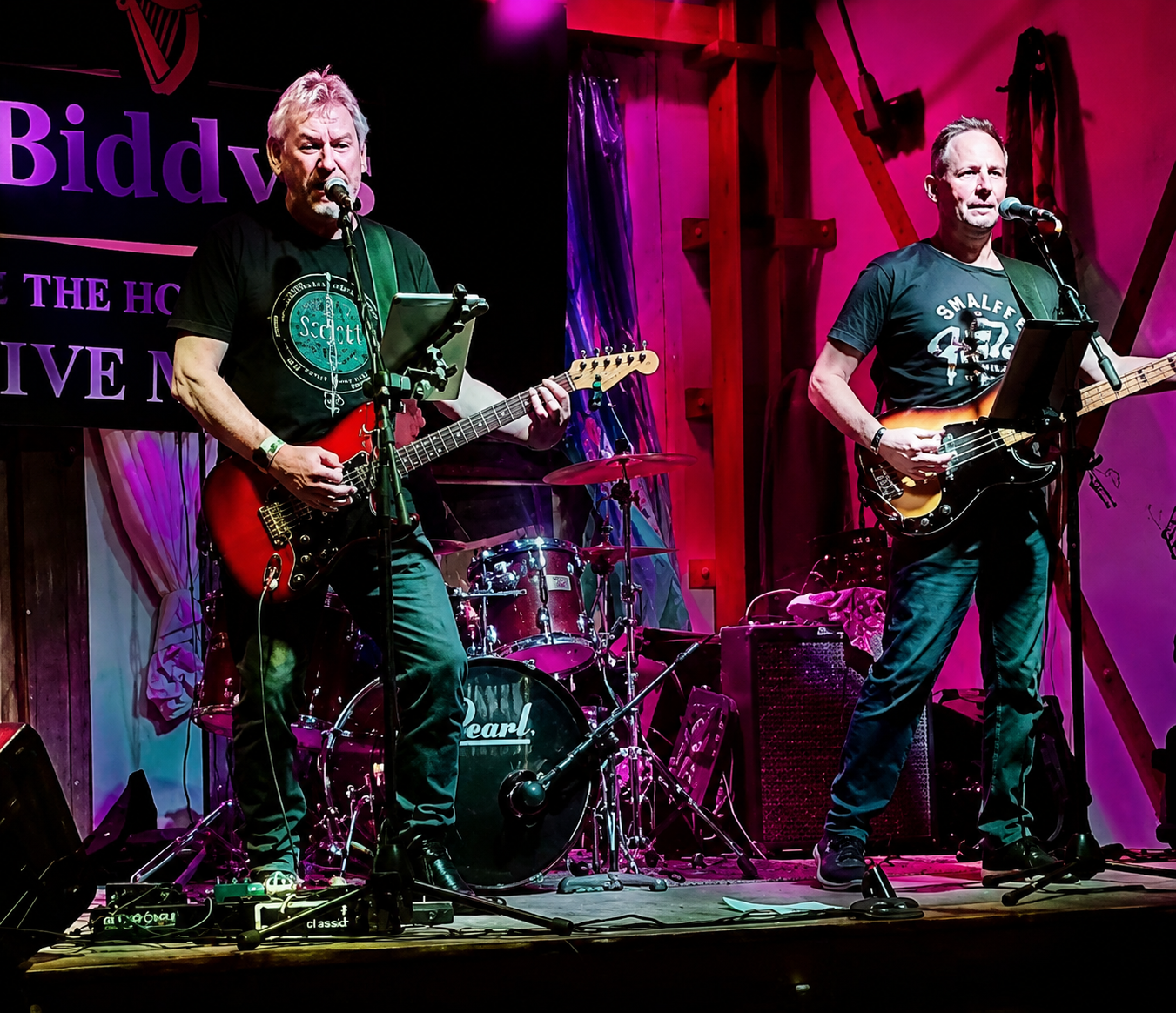 Two male musicians performing on stage with guitars, a drum set in the background, and colorful stage lighting.