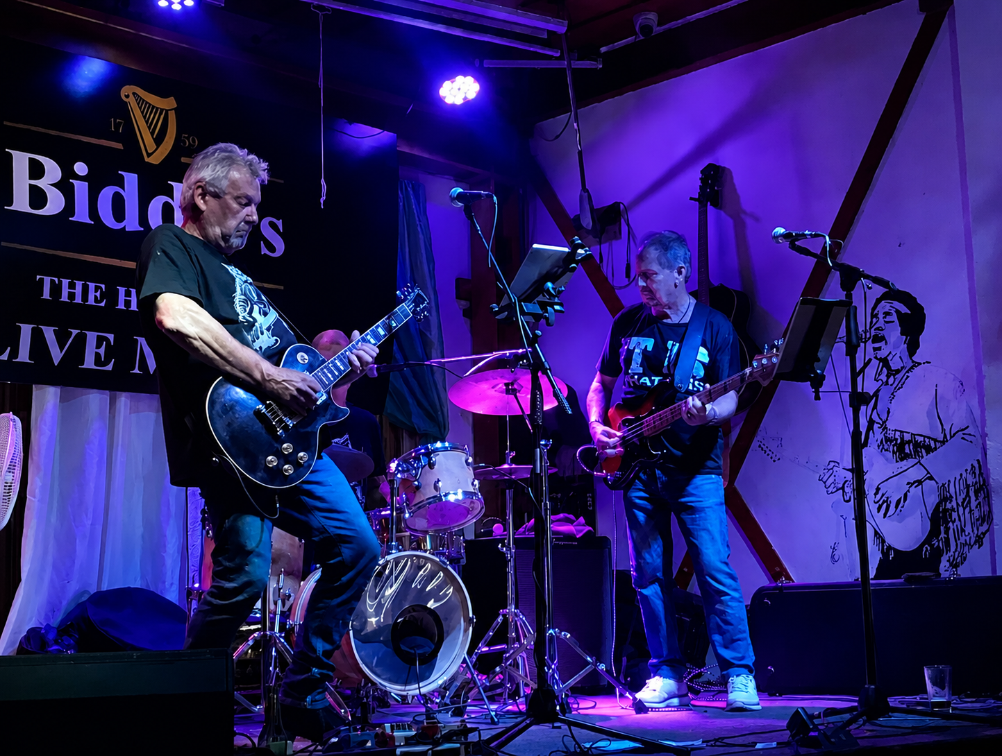 Two musicians performing on stage, one playing an electric guitar and the other playing a bass guitar, with a drum set in the background, in a dimly lit venue with purple and blue lighting.