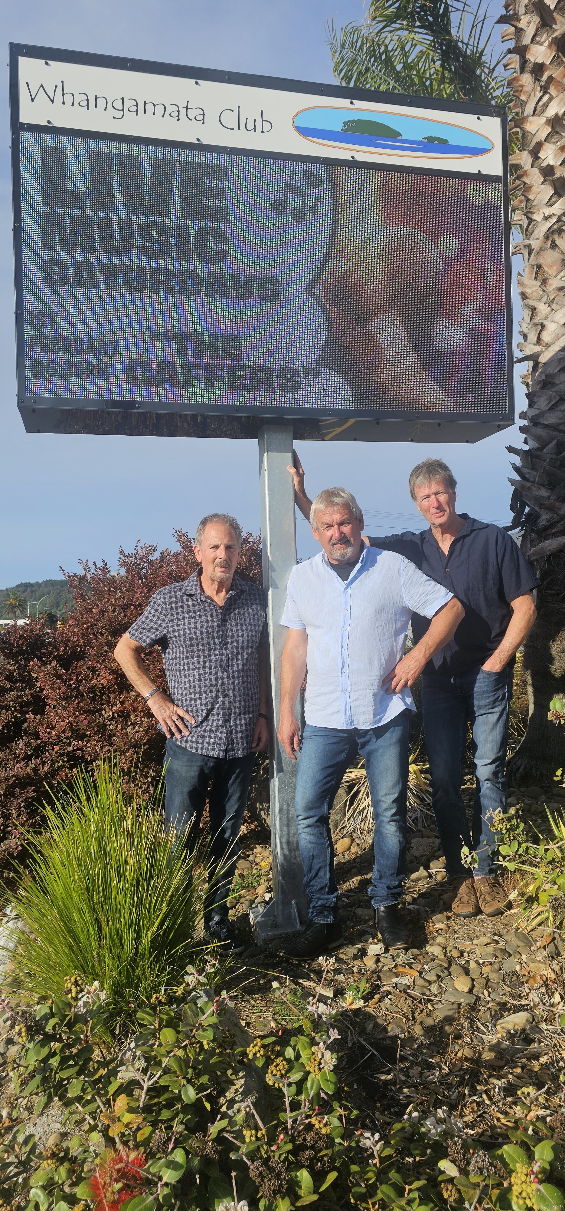 Three men standing outdoors beside a large digital billboard displaying an island graphic and text, with plants and trees in the background.