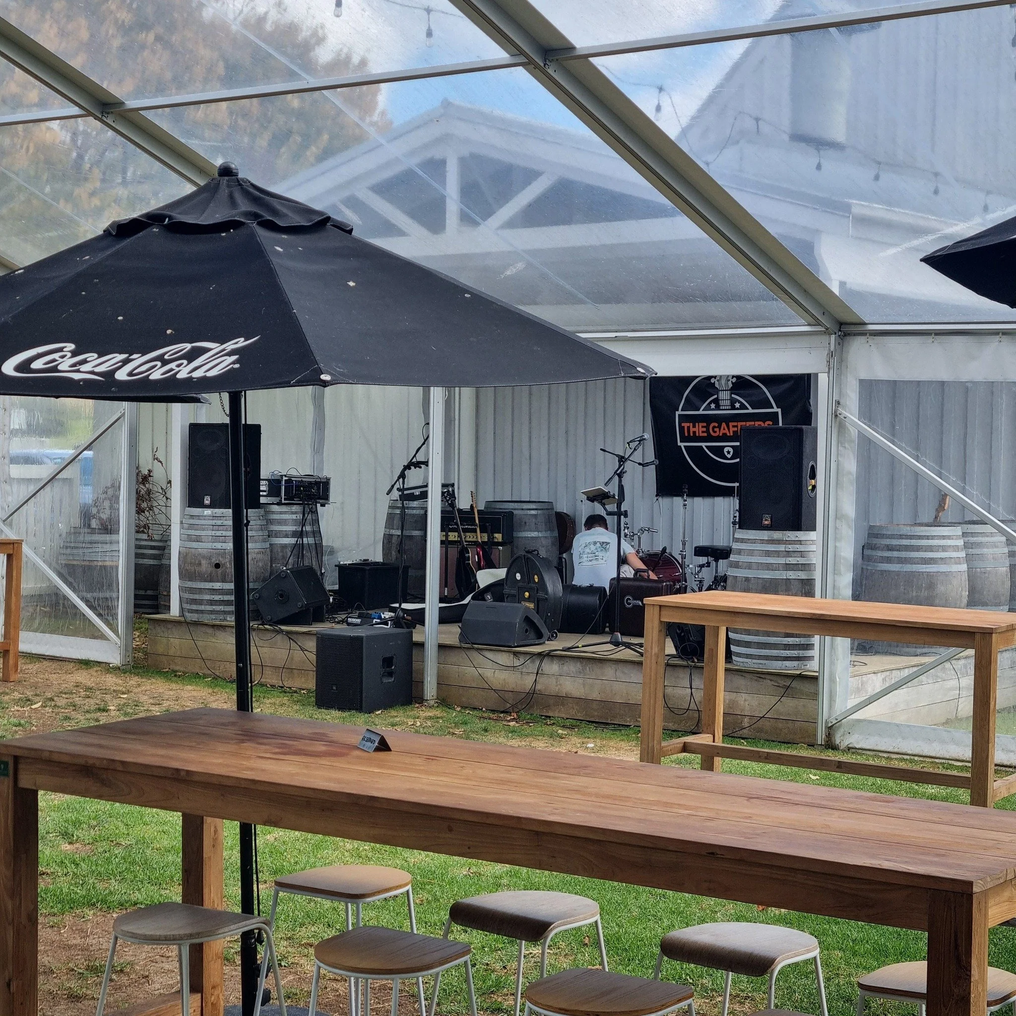 View of an outdoor-stage setup under a clear plastic roof, with musical instruments and speakers, surrounded by tables and stools, in a backyard or garden area.