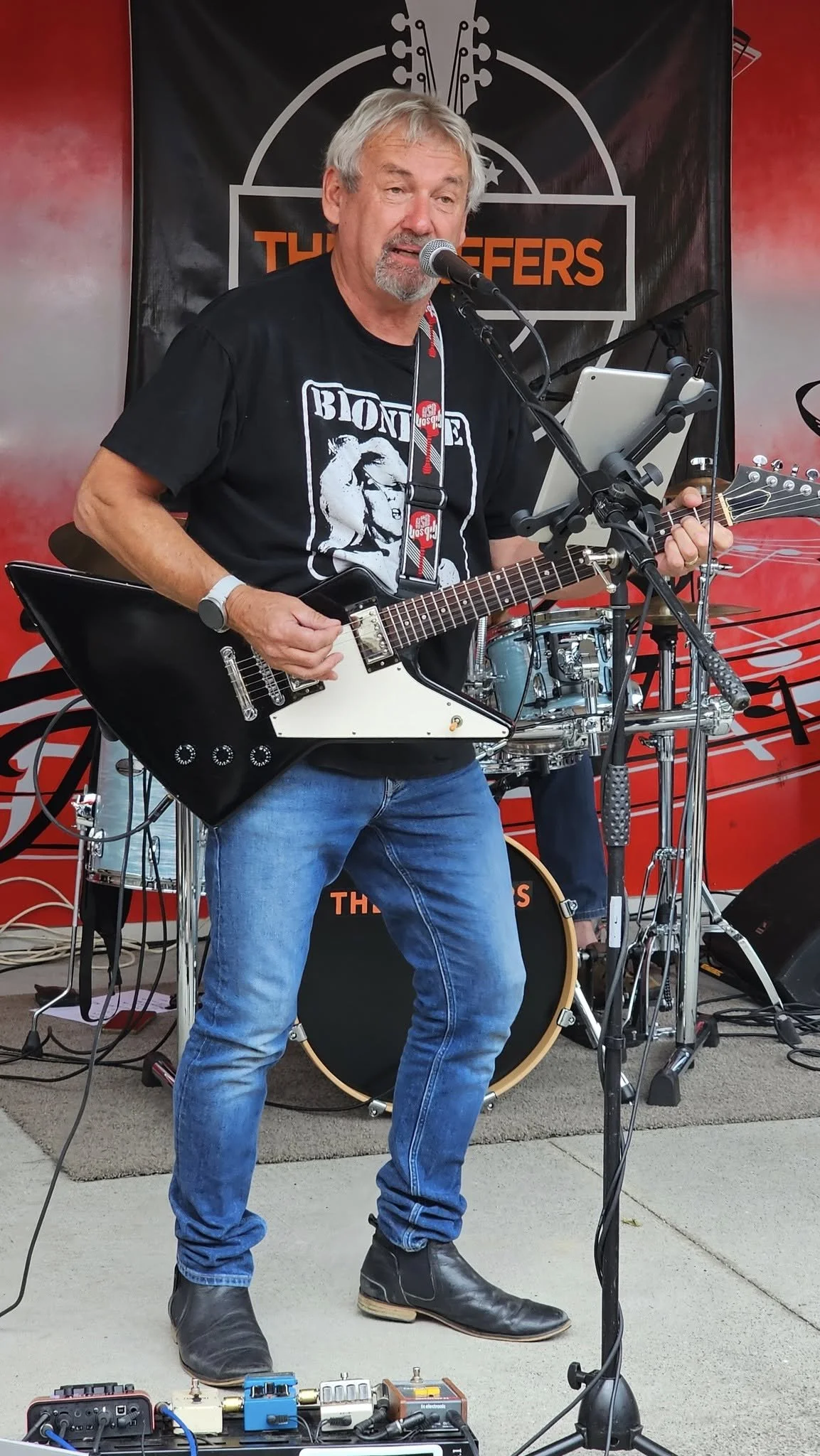 A man with gray hair and a beard singing into a microphone while playing an electric guitar during a live performance at an outdoor event.