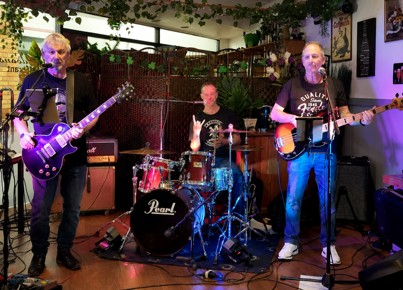 Three musicians performing at a venue. The left musician is playing an electric guitar and singing into a microphone. The middle musician is sitting at a drum set and making a hand gesture. The right musician is playing a bass guitar and singing into