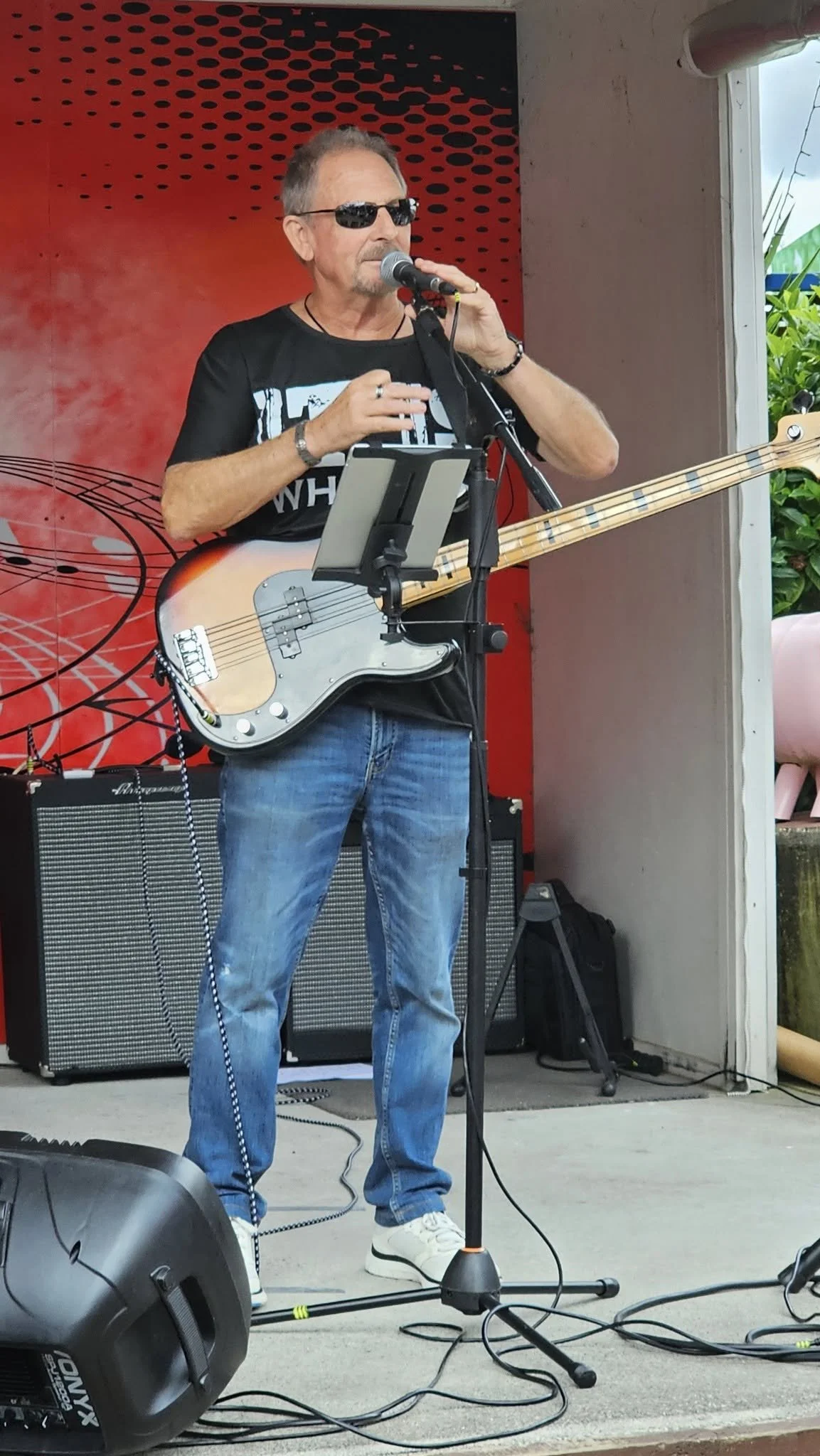 A man with sunglasses, a black T-shirt, and jeans singing into a microphone while playing an electric bass guitar on stage, with music equipment in the background.