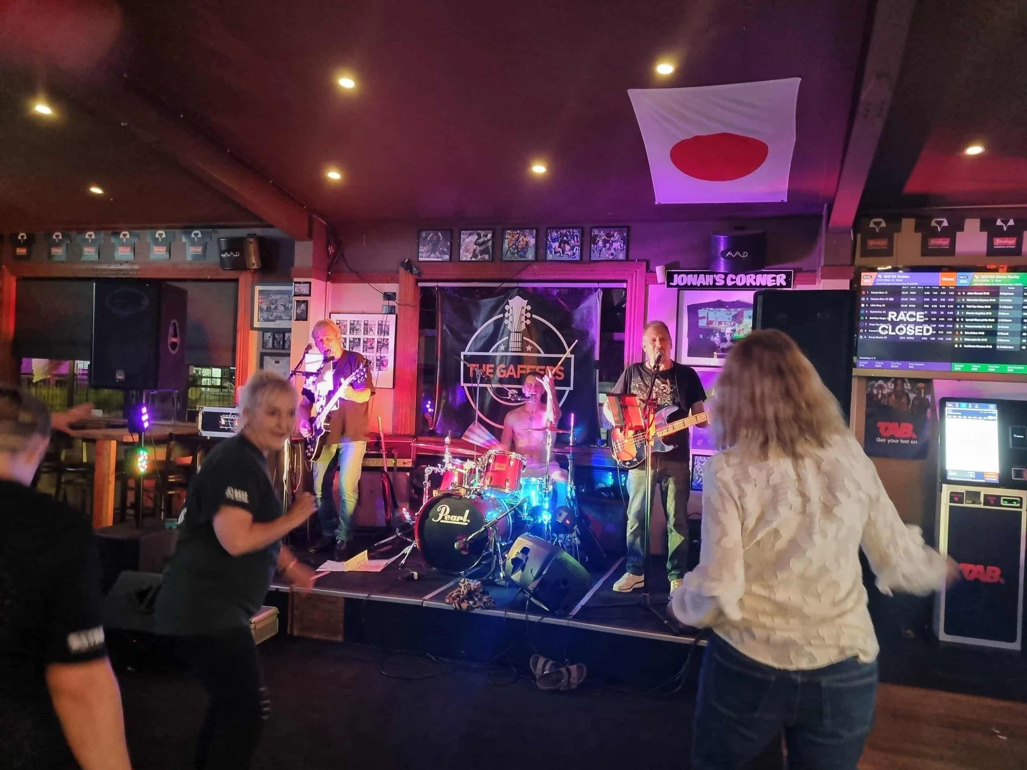 Live music performance at an indoor bar or club featuring a band with a guitarist, bassist, and drummer, with audience members dancing and enjoying the show, Japanese flag hanging on the ceiling, and colorful lights creating a vibrant atmosphere.