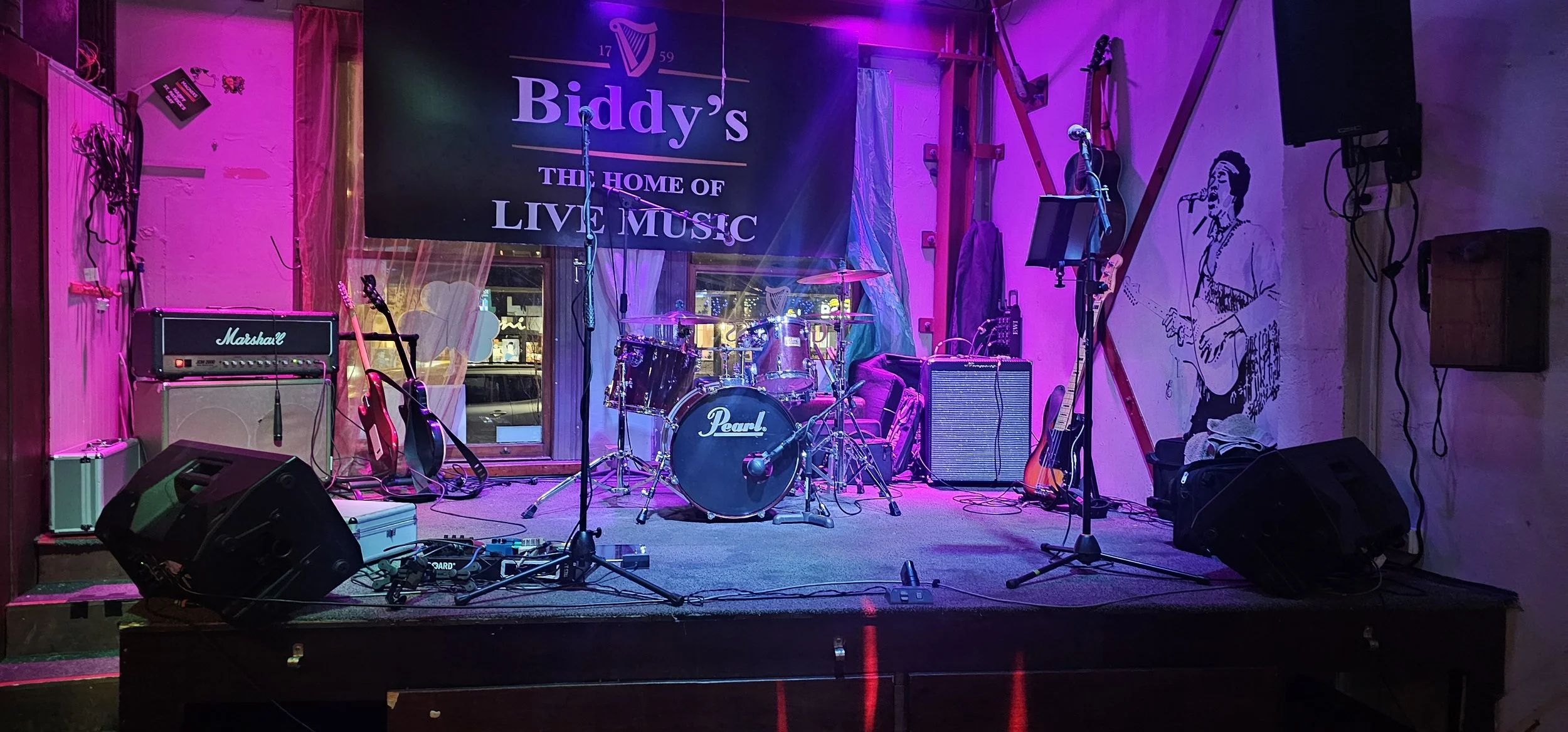 Empty stage set up for live music performance with musical instruments including drums, guitars, and amplifiers, under purple lighting, with a sign behind reading "Biddy's The Home of Live Music".