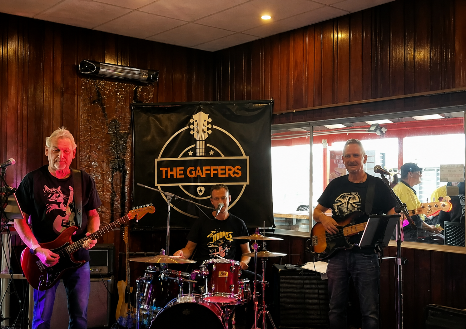 A band called The Gaffers performing on stage with three musicians playing guitar and drums, inside a venue with wooden walls and a large window.