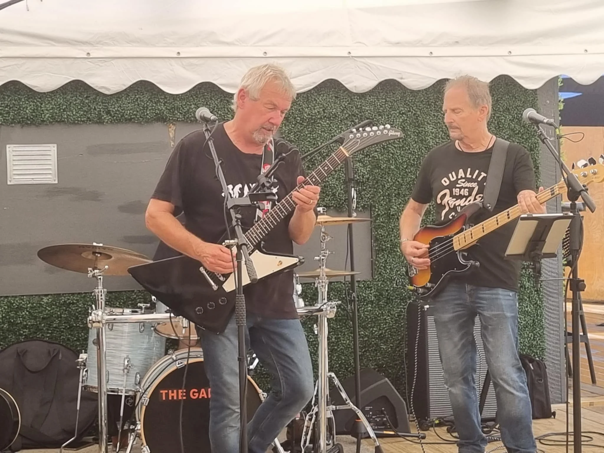 Two men playing guitars on a stage under a white tent, with musical equipment behind them.