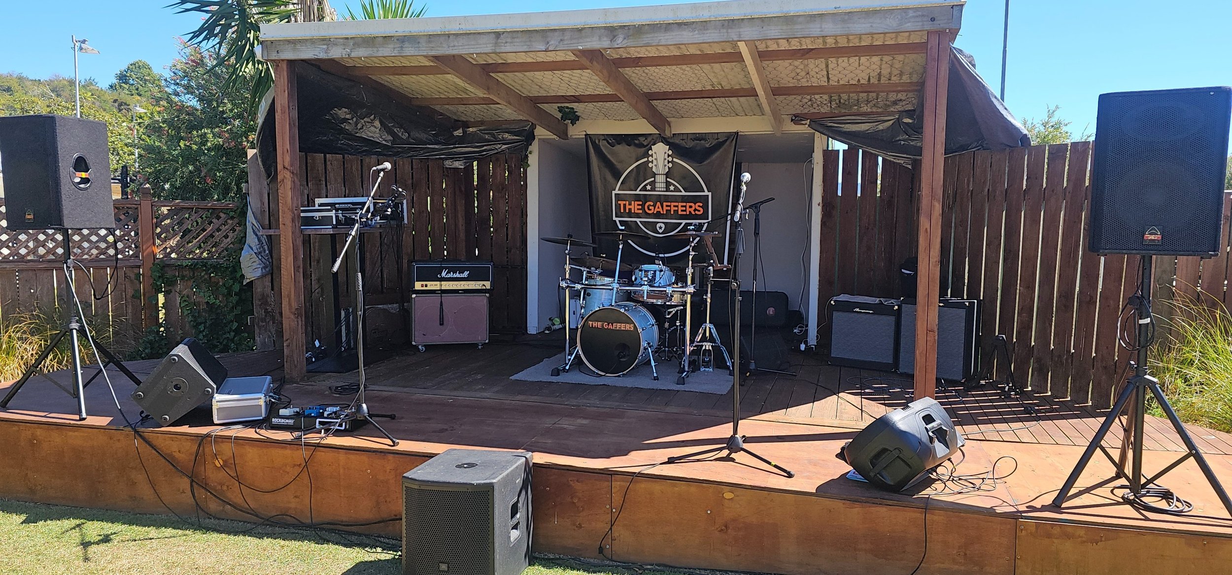An outdoor music stage set up with various instruments including drums, guitars, and microphones. Large speakers and audio equipment are positioned on either side of the stage, which has a wooden platform and a roof. A banner displaying the band name