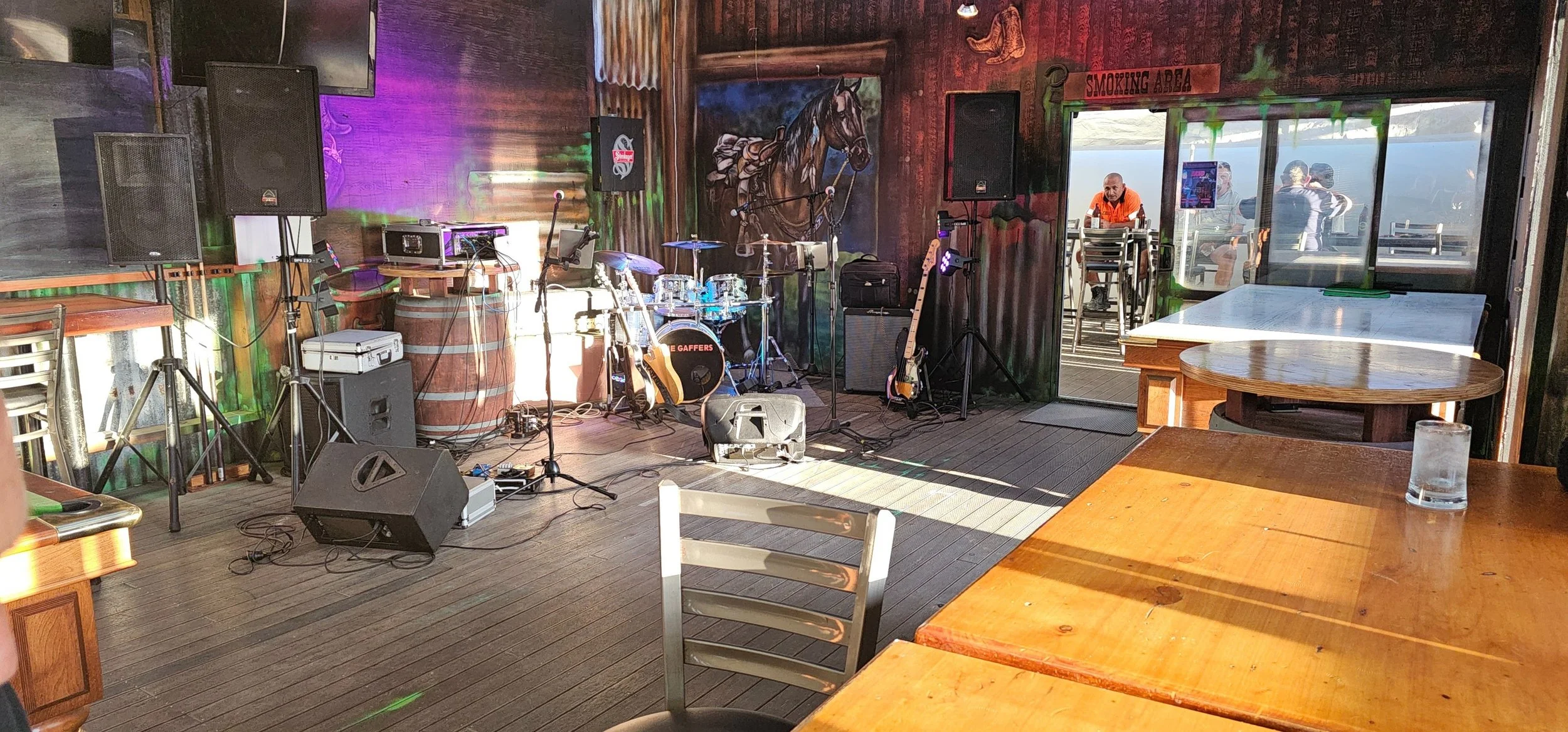 Indoor stage area with musical instruments including drums, guitars, and microphones, set up for a live performance inside a rustic venue with wooden walls, tables, and barstools, and a few people visible outside through large windows.
