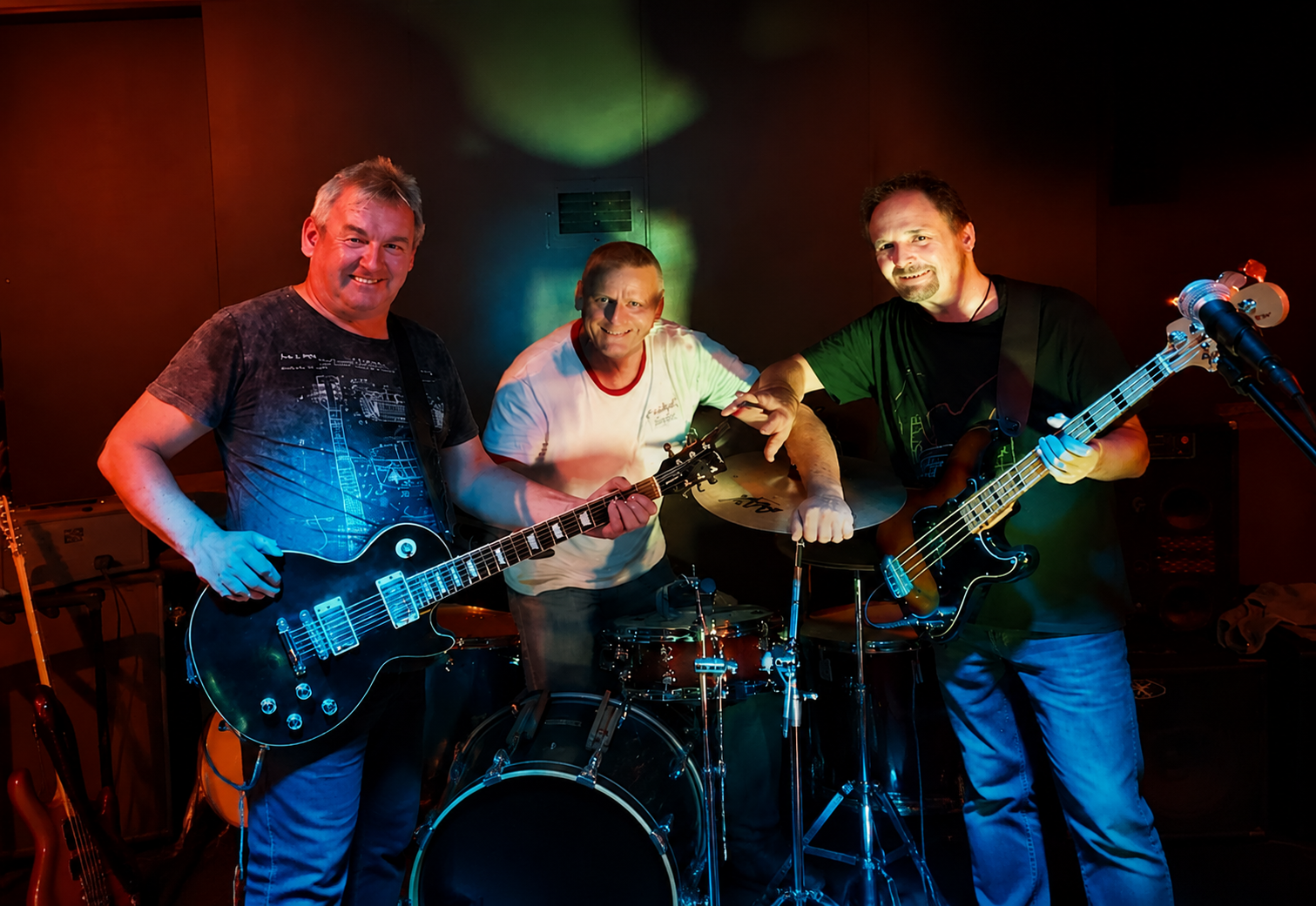 Three men in a band with guitars and drums on stage, smiling and posing for the photo under colorful stage lights.