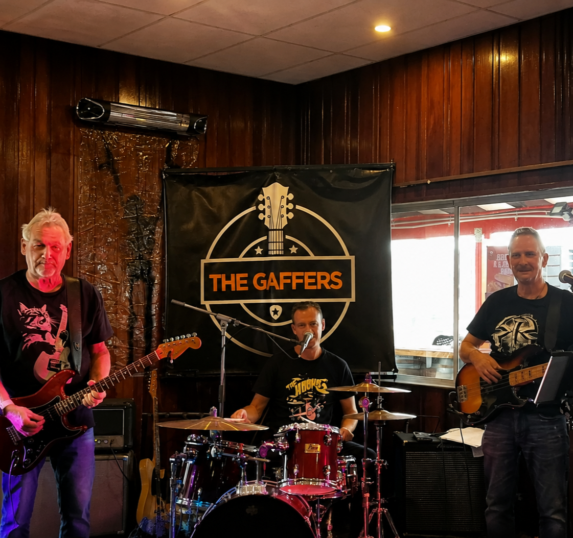 A three-piece band performing live on stage at a venue called The Gaffers, with a wooden decor background and a large sign behind them. The band includes a guitarist on the left, a drummer in the center, and a bassist on the right.