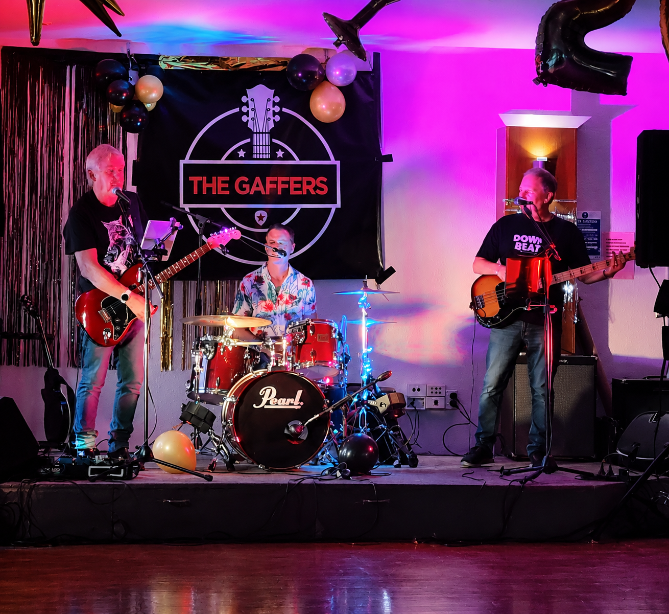 Three musicians performing on stage with a black banner reading 'The Gaffers' in the background, decorated with balloons and shiny streamers, under colorful stage lighting.