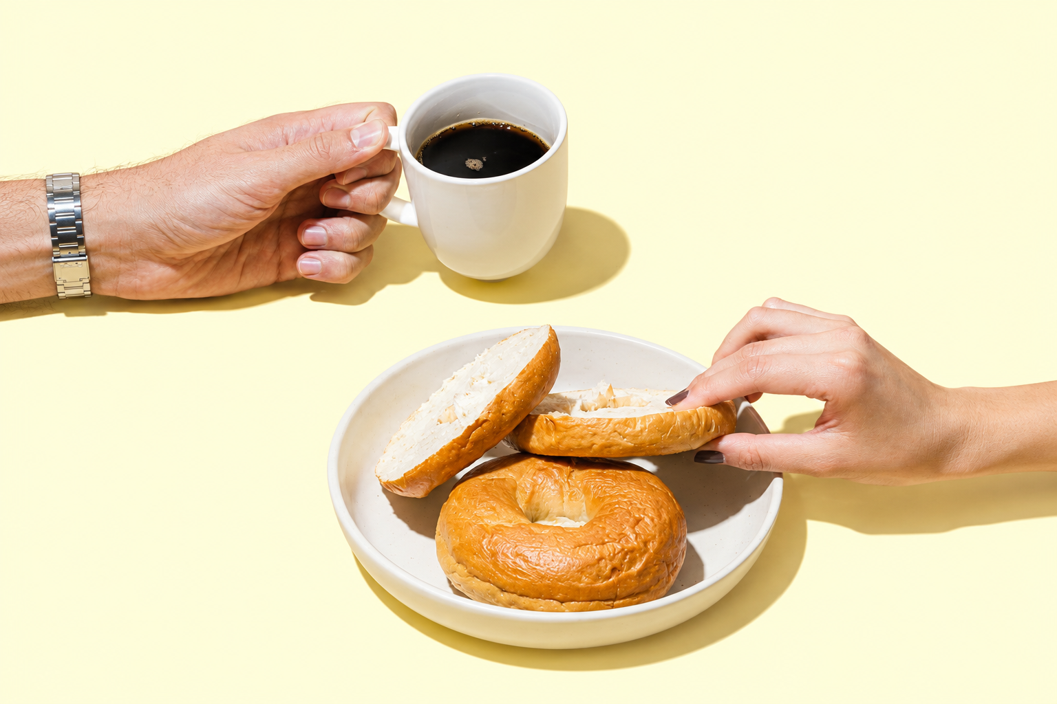 A person holding a white mug filled with black coffee, and another person reaching for a bagel with cream cheese on a white plate, on a light yellow background.