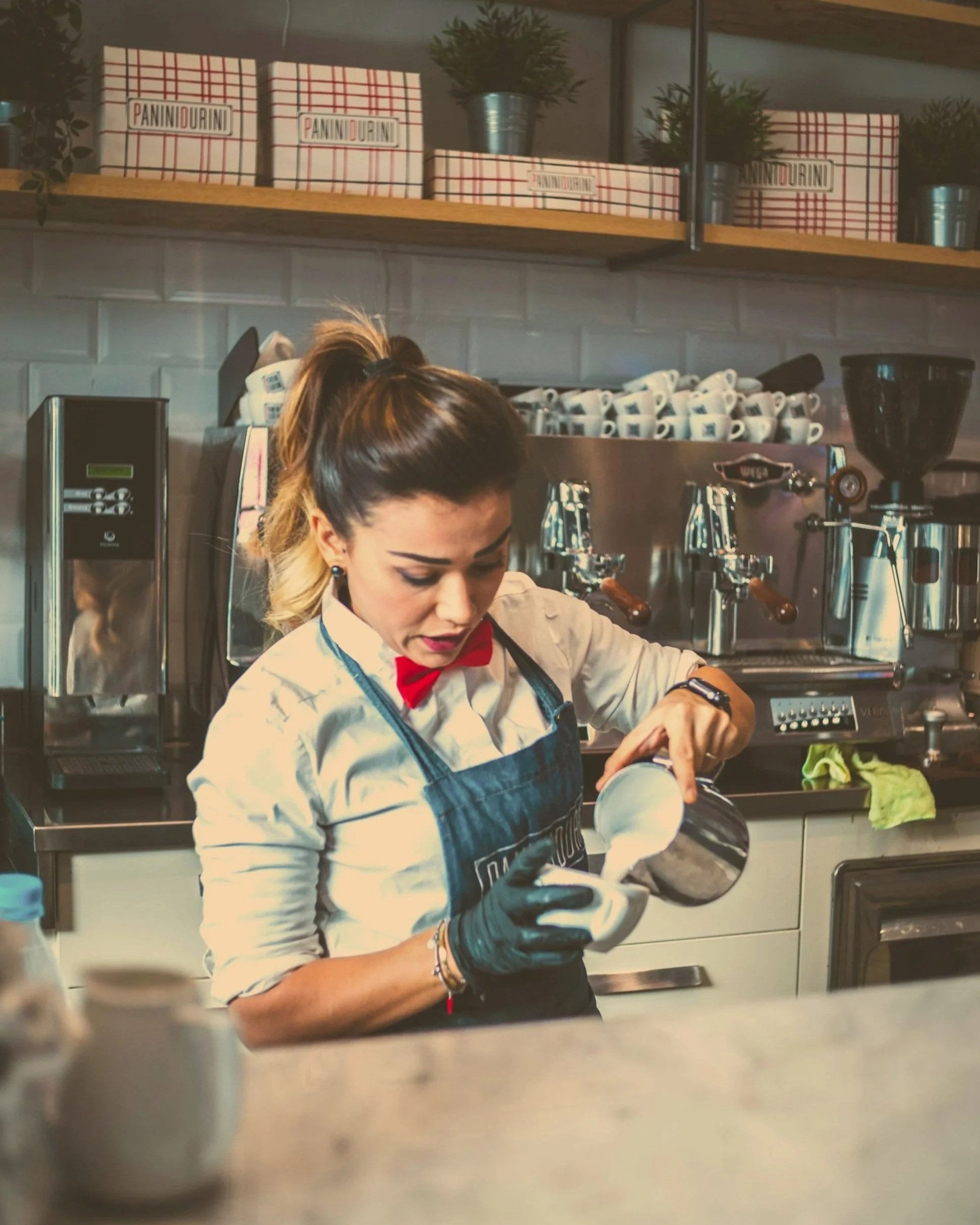A woman with blonde hair tied back, wearing a white shirt, black gloves, a red bow tie, and an apron, is preparing a latte art in a coffee shop kitchen with coffee machines, cups, and boxes with the word "Panini Durini" on shelves in the background.