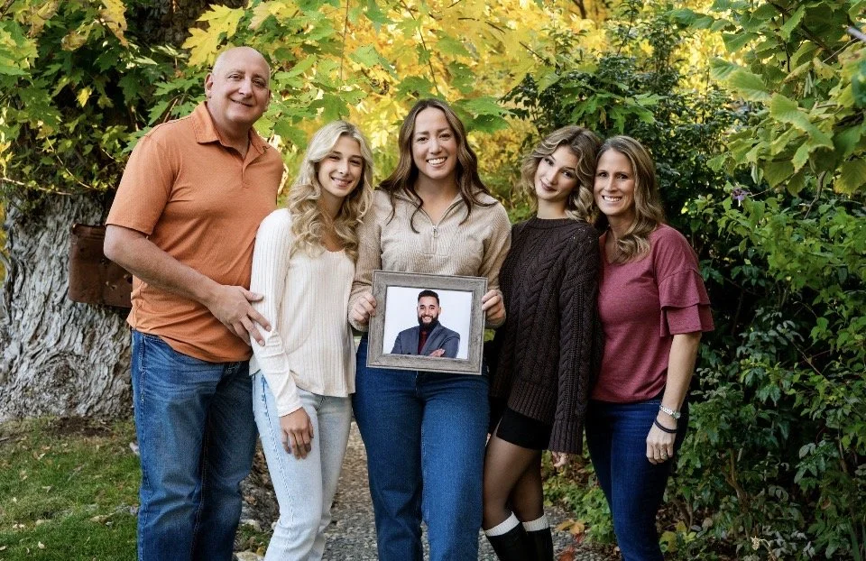 Group of five people standing outdoors in front of trees with yellow and green leaves, with one person holding a framed photo of a man.