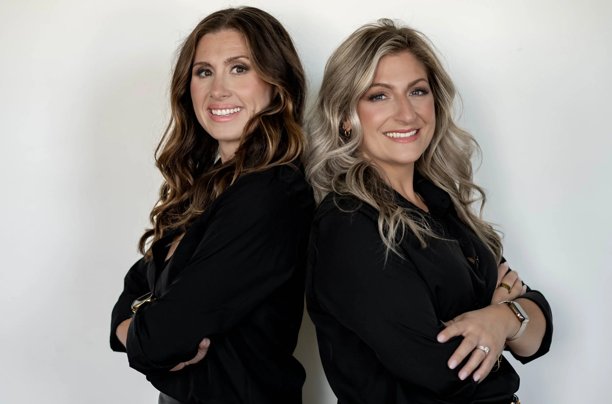 Two women standing back-to-back, smiling, with arms crossed, against a plain white background.
