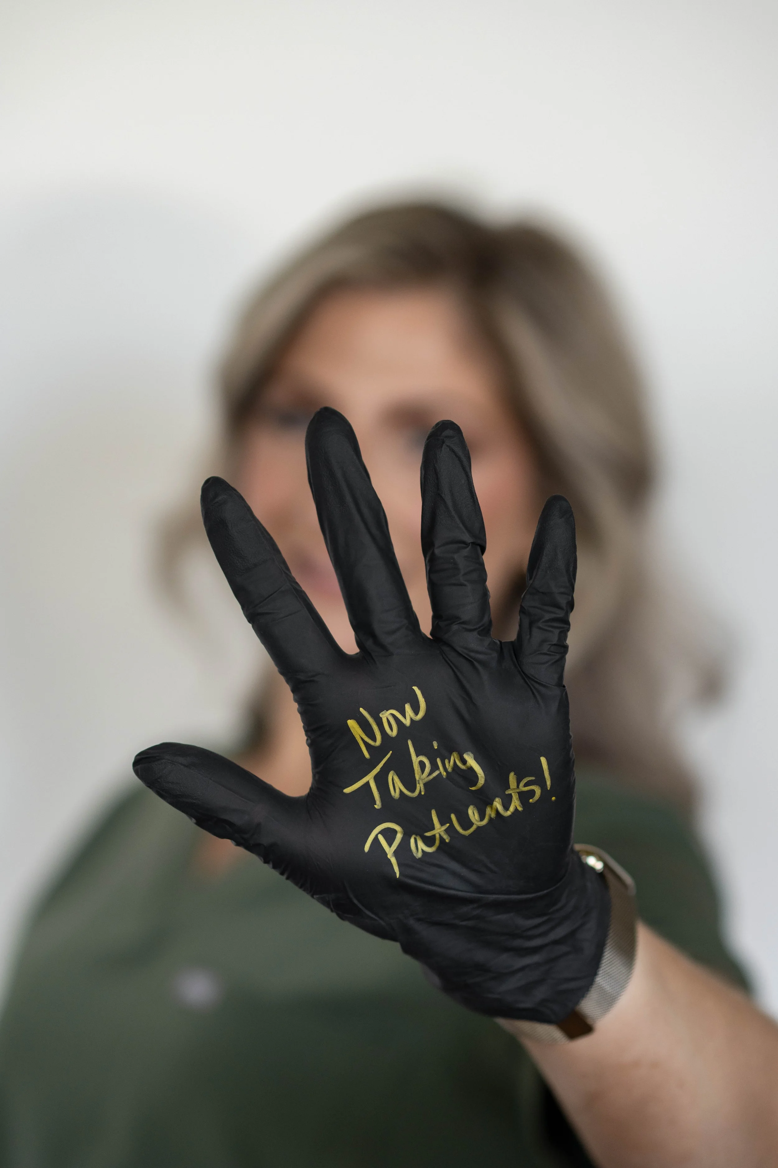 A woman with short, gray hair wears a black glove with gold writing that says "Now Taking Patients!" and is holding up her hand in front of her face, which is out of focus.
