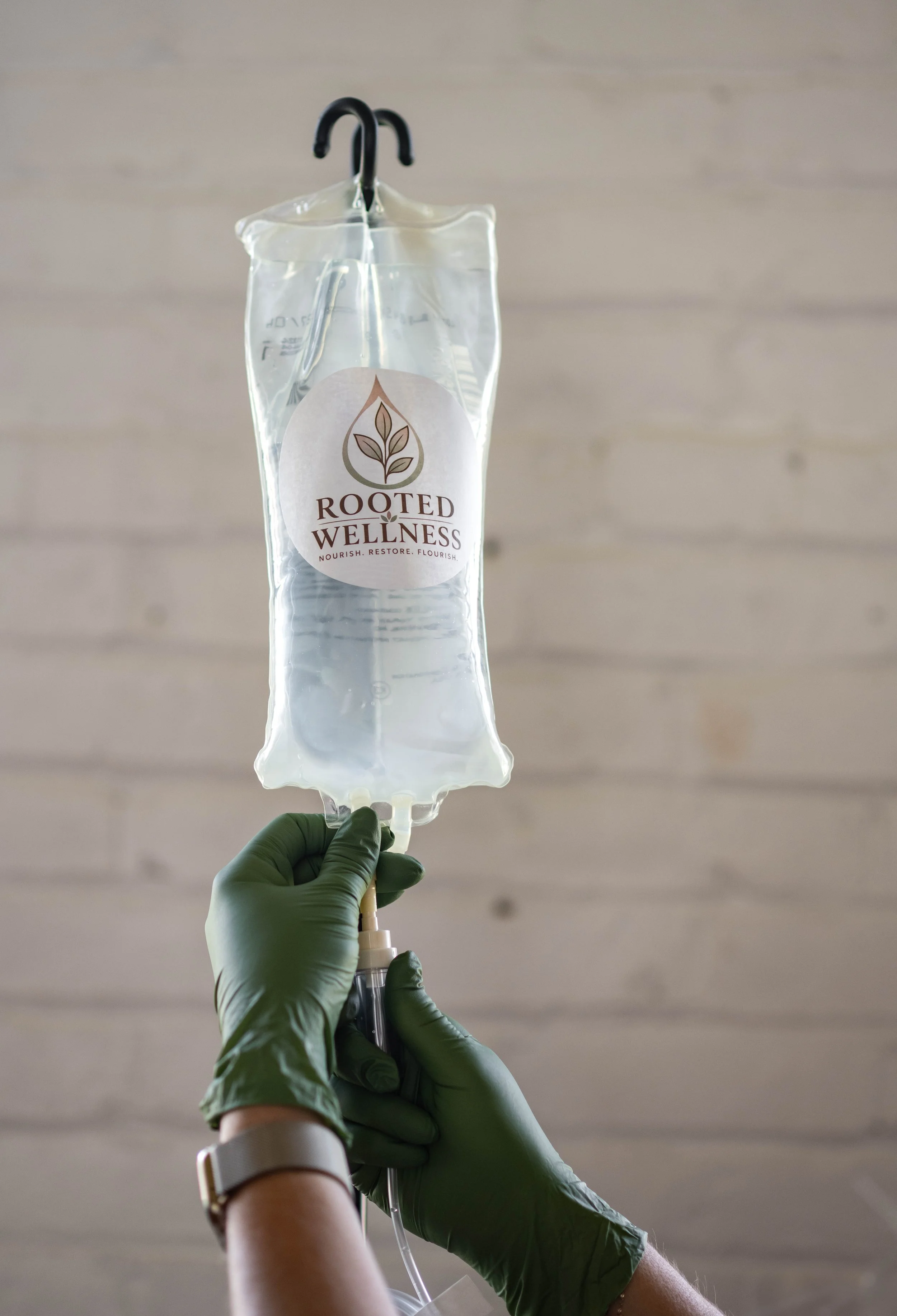 A person wearing green gloves and a watch is holding an IV fluid bag with a logo that reads 'Rooted Wellness' and a leaf symbol, hanging from a black hook.