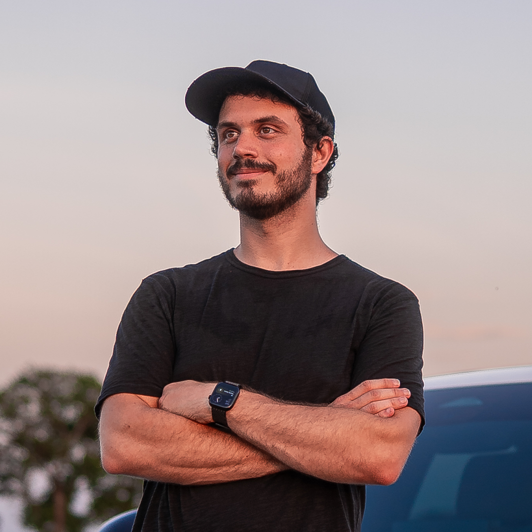 A young man with a beard and curly hair, wearing a black cap, black t-shirt, and smartwatch, standing outdoors during sunset with his arms crossed and smiling.
